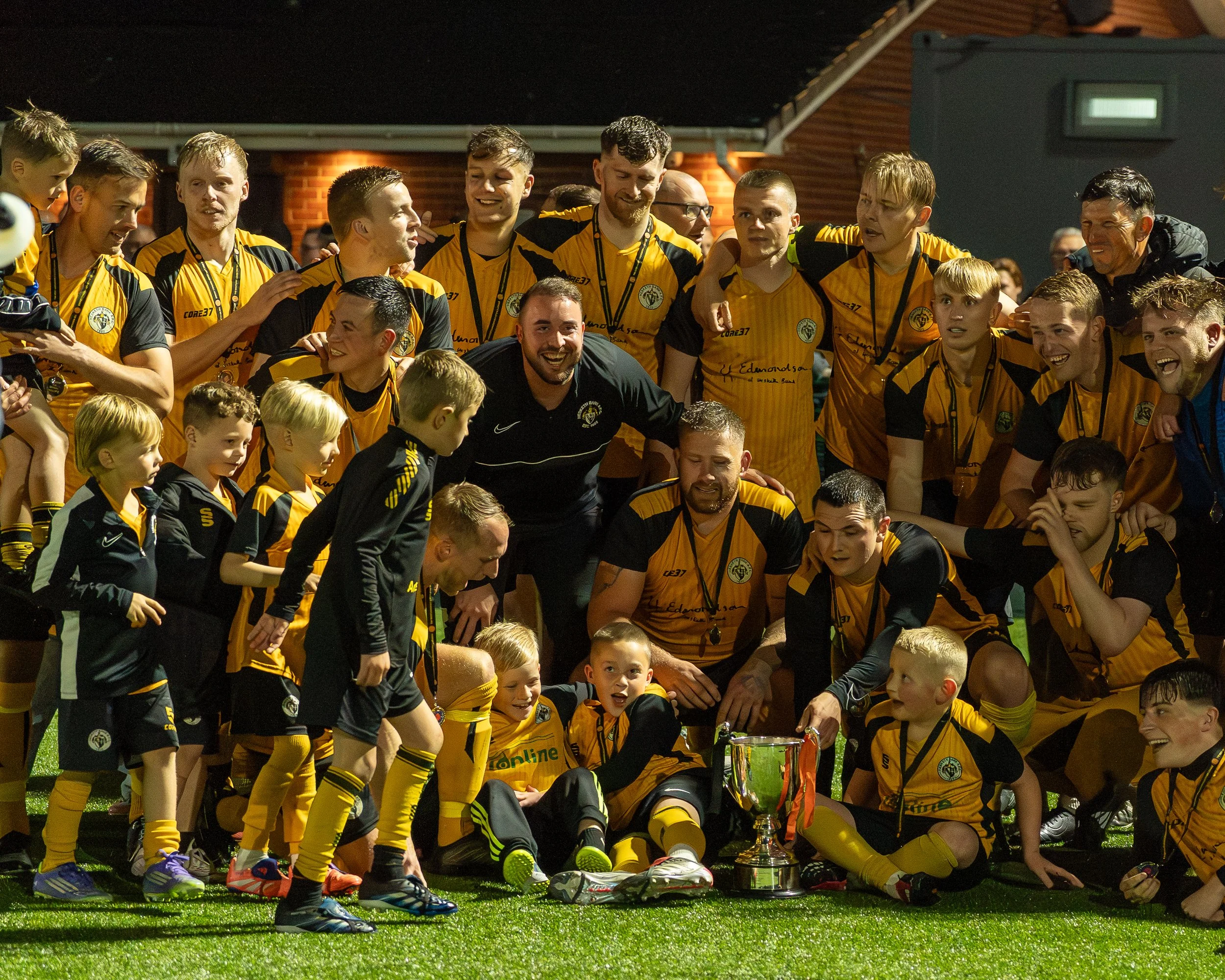 Group of soccer players in yellow and black uniforms celebrating with medals and a large trophy.