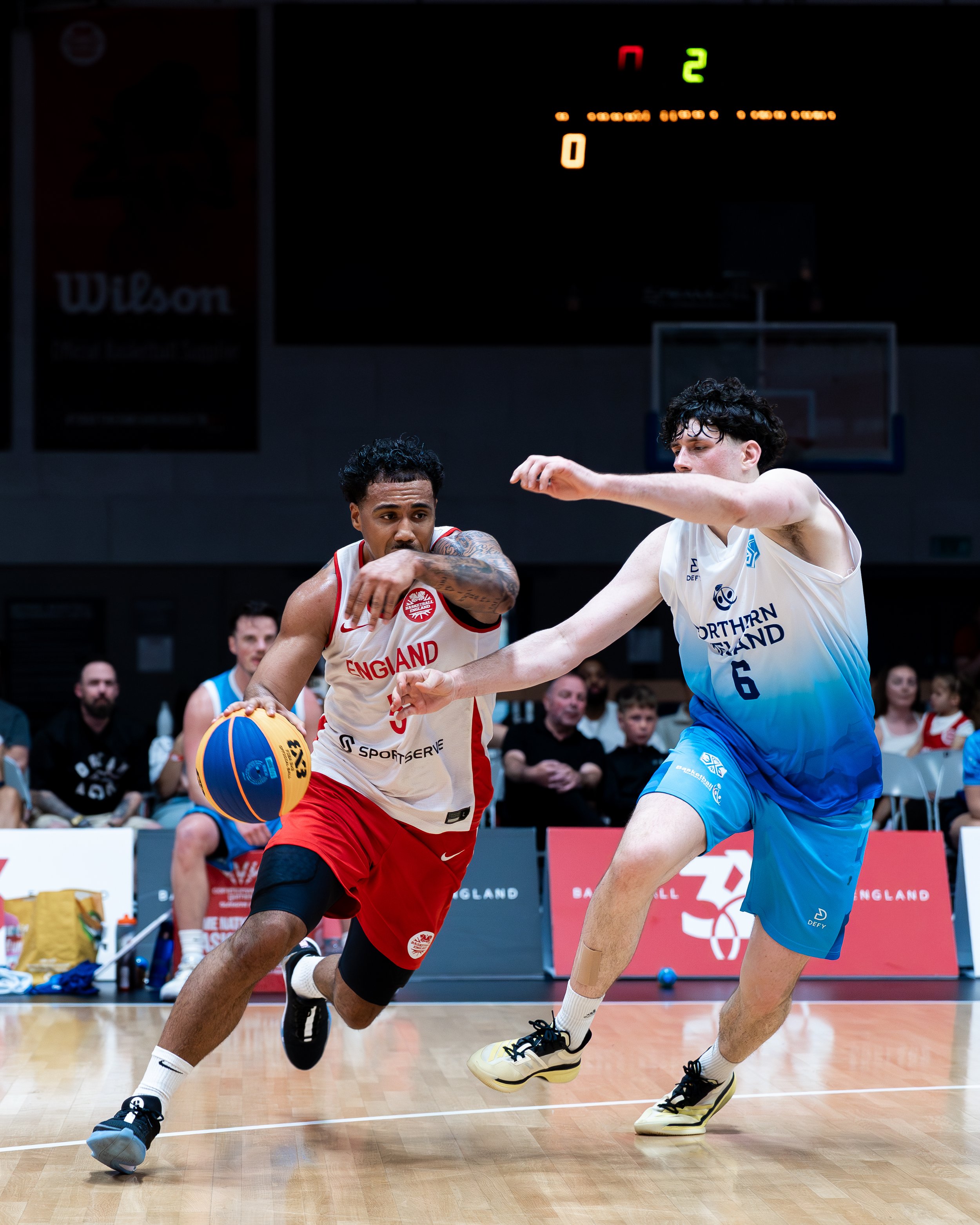 Two basketball players from opposing teams competing for control of a basketball during a game; one from England in a white and red uniform, the other from Northern Ireland in a blue and white uniform.