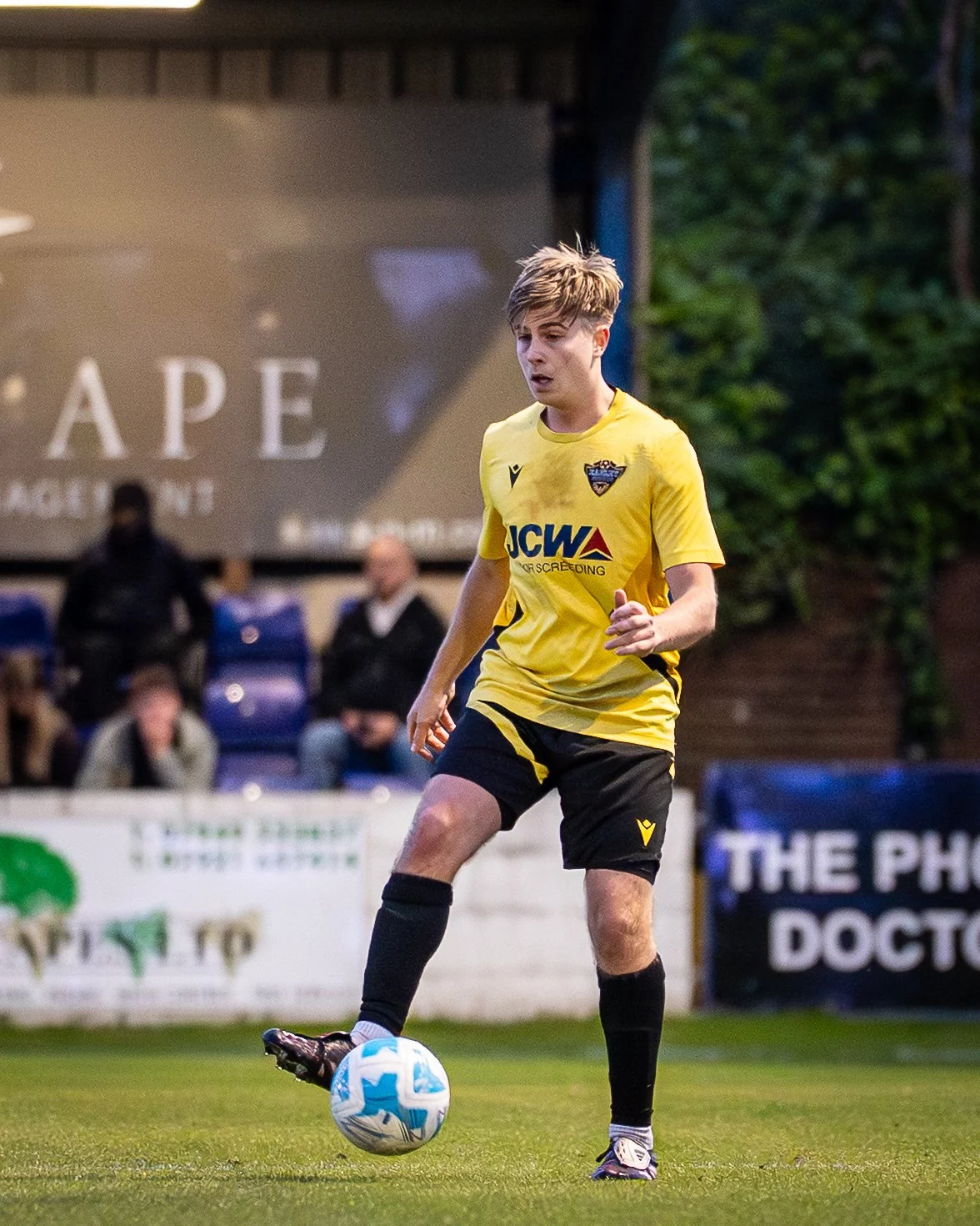 A young male soccer player in a yellow jersey with black shorts and socks is on a soccer field, controlling a white and blue soccer ball.