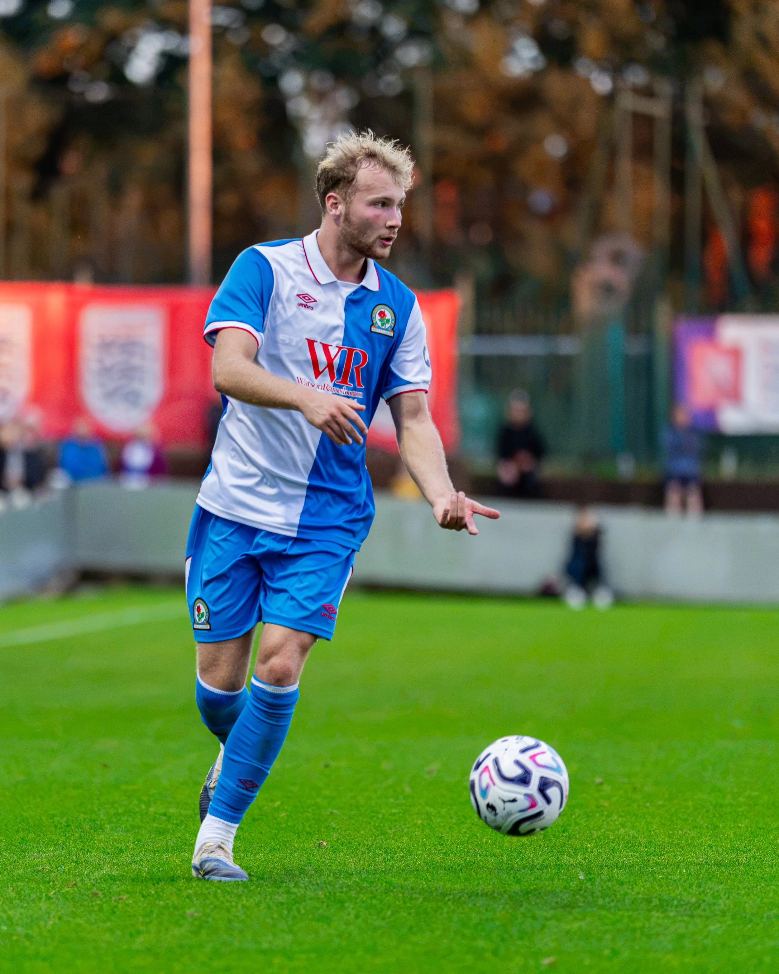 A male soccer player in a blue and white uniform is on a soccer field, about to kick a soccer ball. The background shows trees and some blurred spectators or structures.