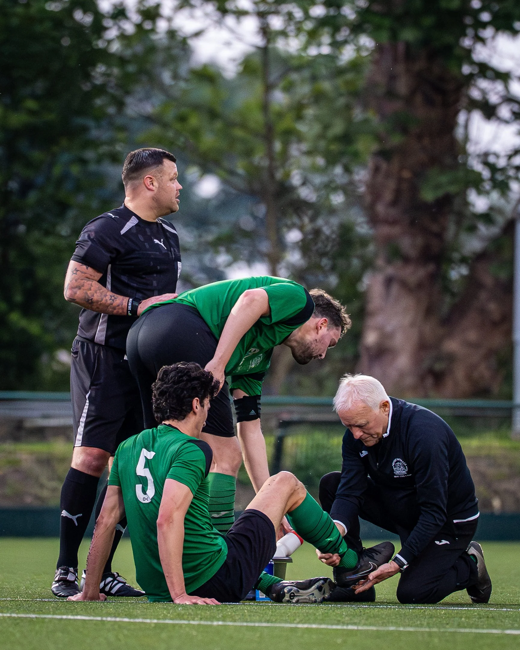A soccer match injury scene with players and a referee. One player in a green jersey is on his knees, being attended to by a man in a black uniform, possibly a medic. Two other players, also in green jerseys, are standing nearby. The background shows
