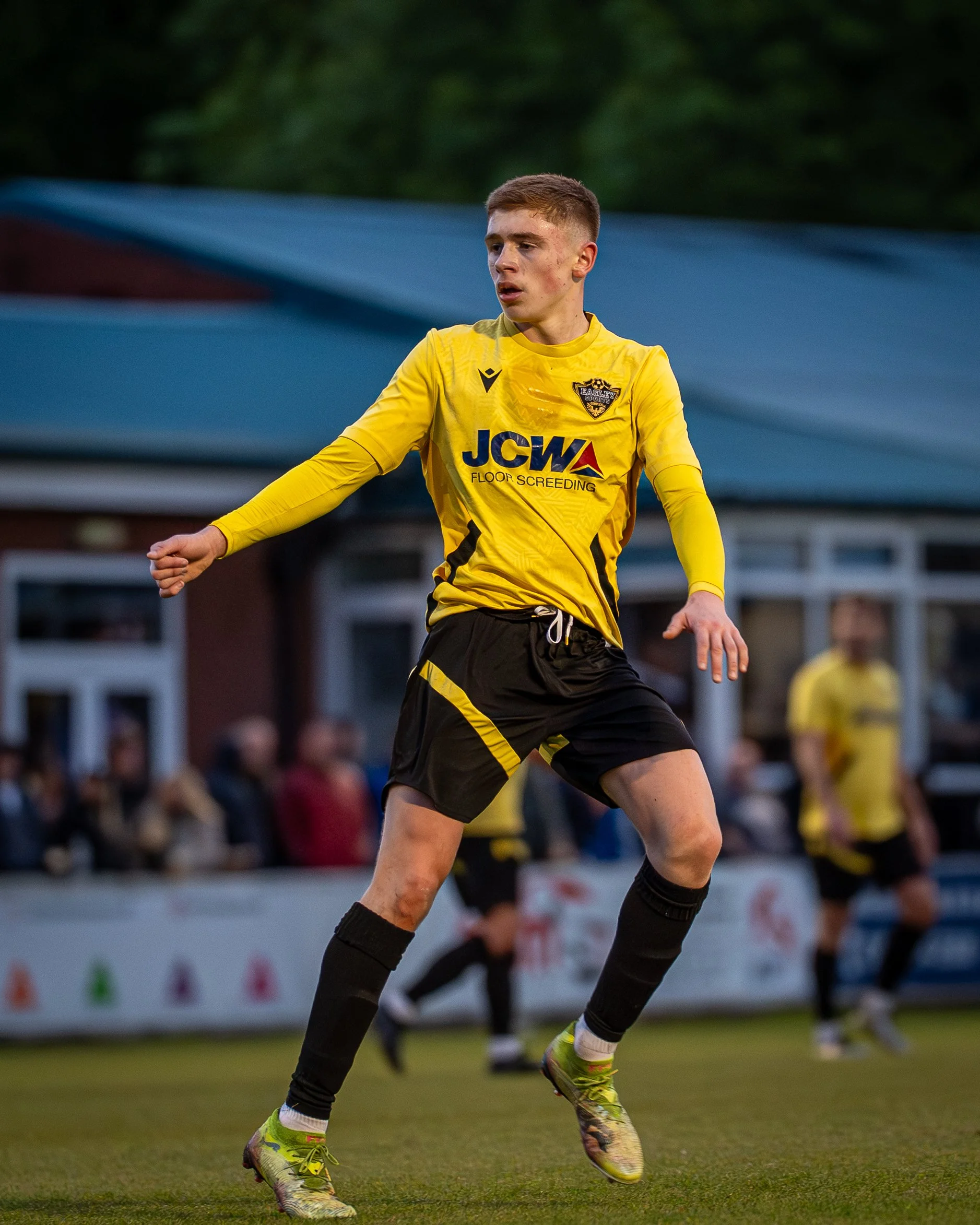 A young male soccer player in a yellow jersey and black shorts on a grassy field, appearing to be in motion during a game or practice, with spectators and structures blurred in the background.