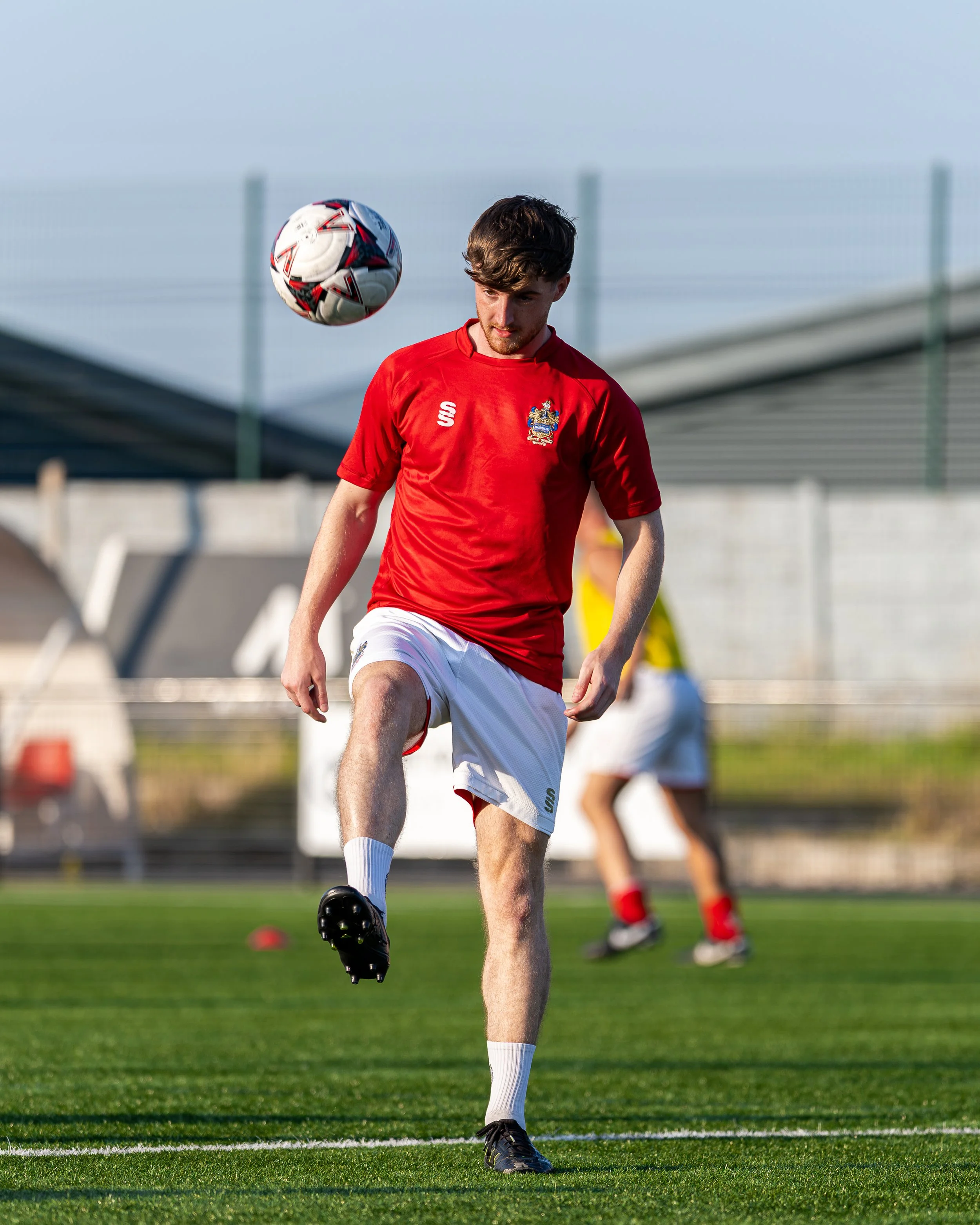A male soccer player in a red jersey and white shorts is kicking a soccer ball on a grass field during practice or game.