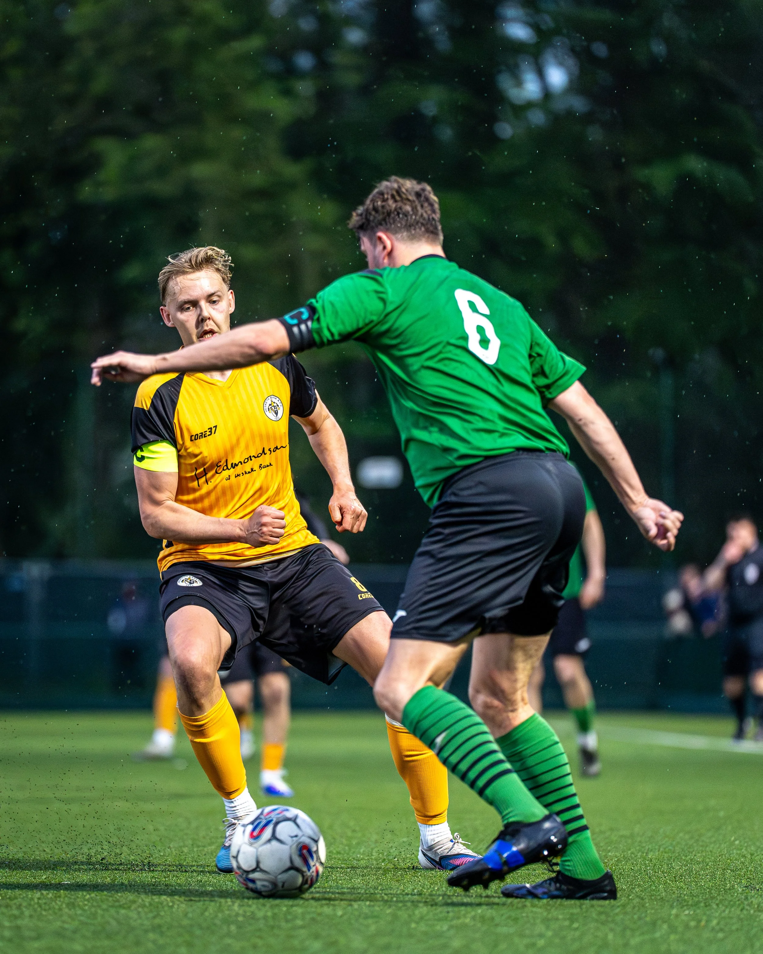 Two soccer players competing for the ball on a green field, one wearing a yellow jersey and the other in a green jersey, with a soccer ball at their feet, and other players blurred in the background.