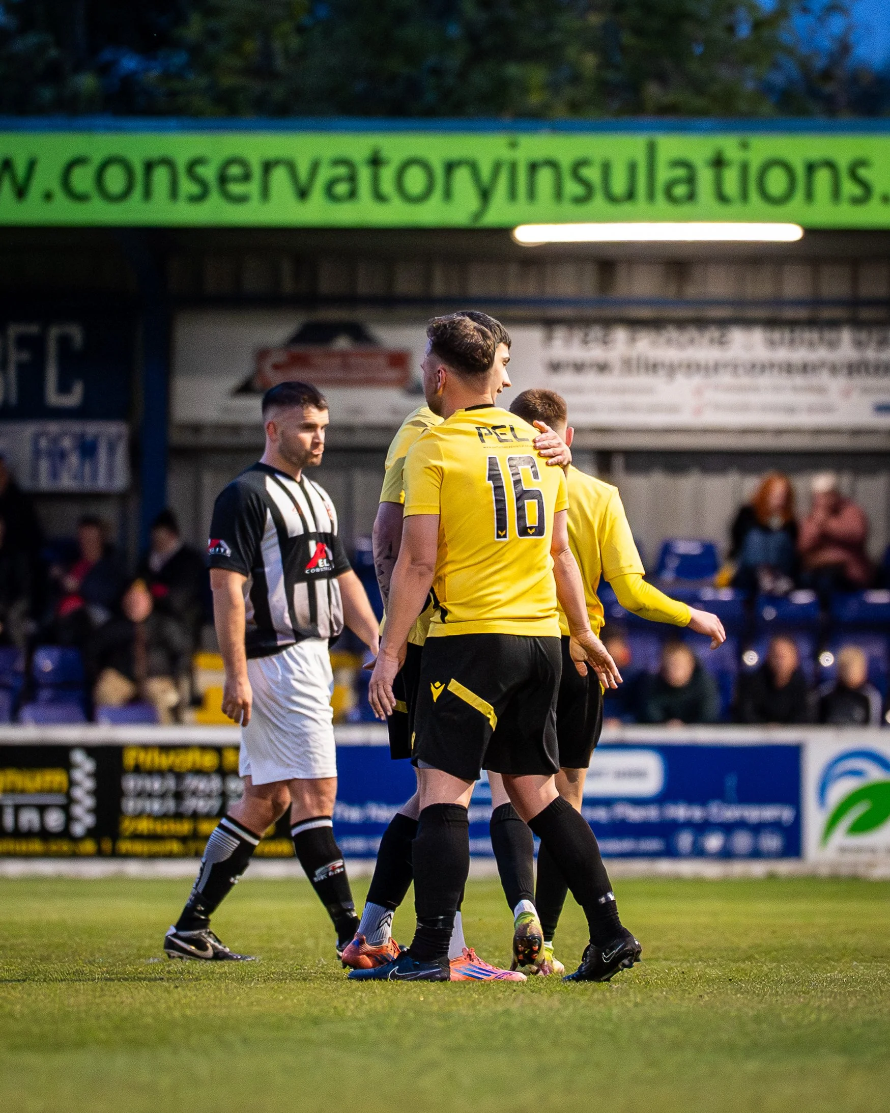 Soccer players in yellow and black uniforms standing on a soccer field during a game, with spectators in the background and advertising signs behind them.