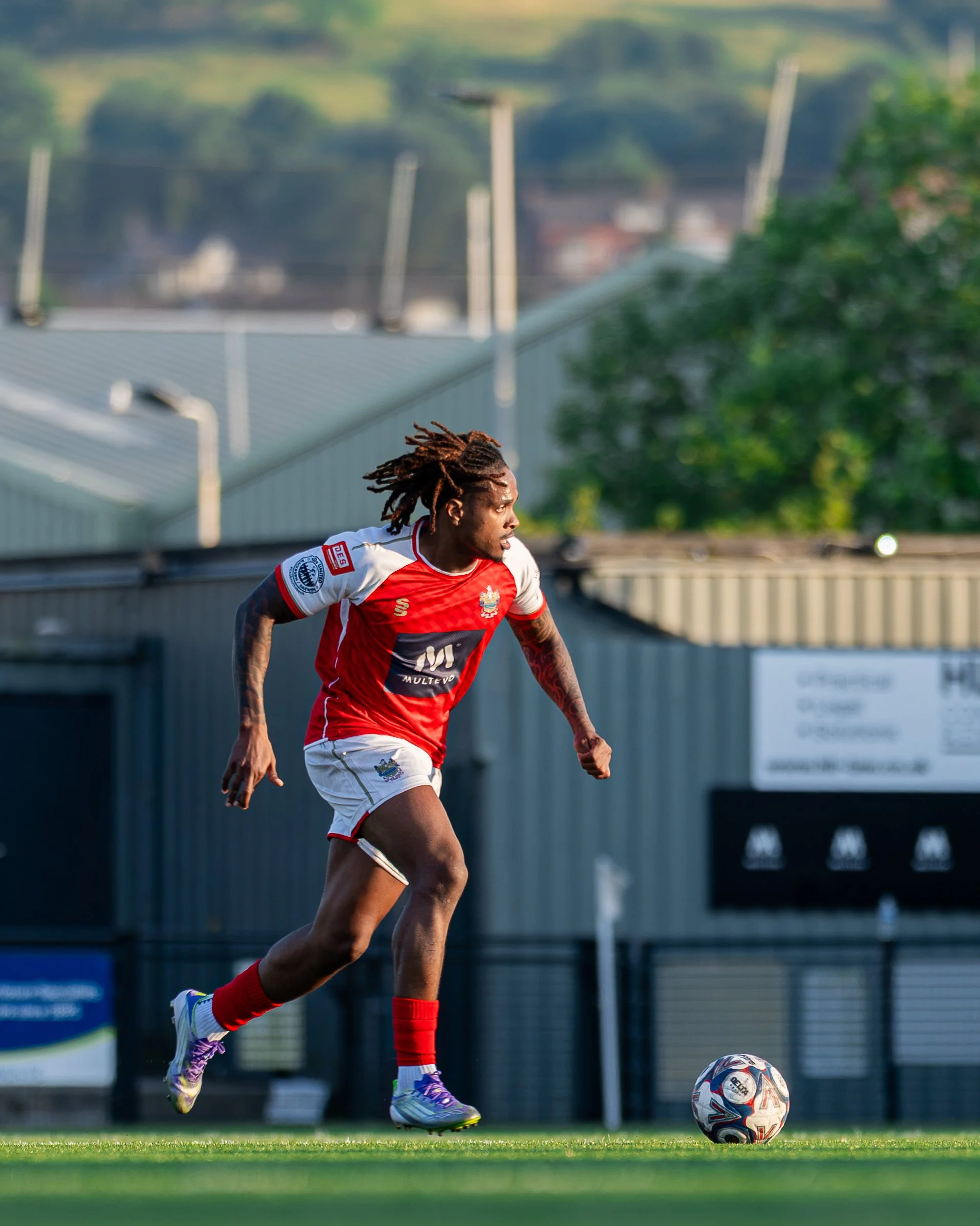 Soccer player with dreadlocks wearing a red and white jersey, running on a soccer field with a ball, during daylight.