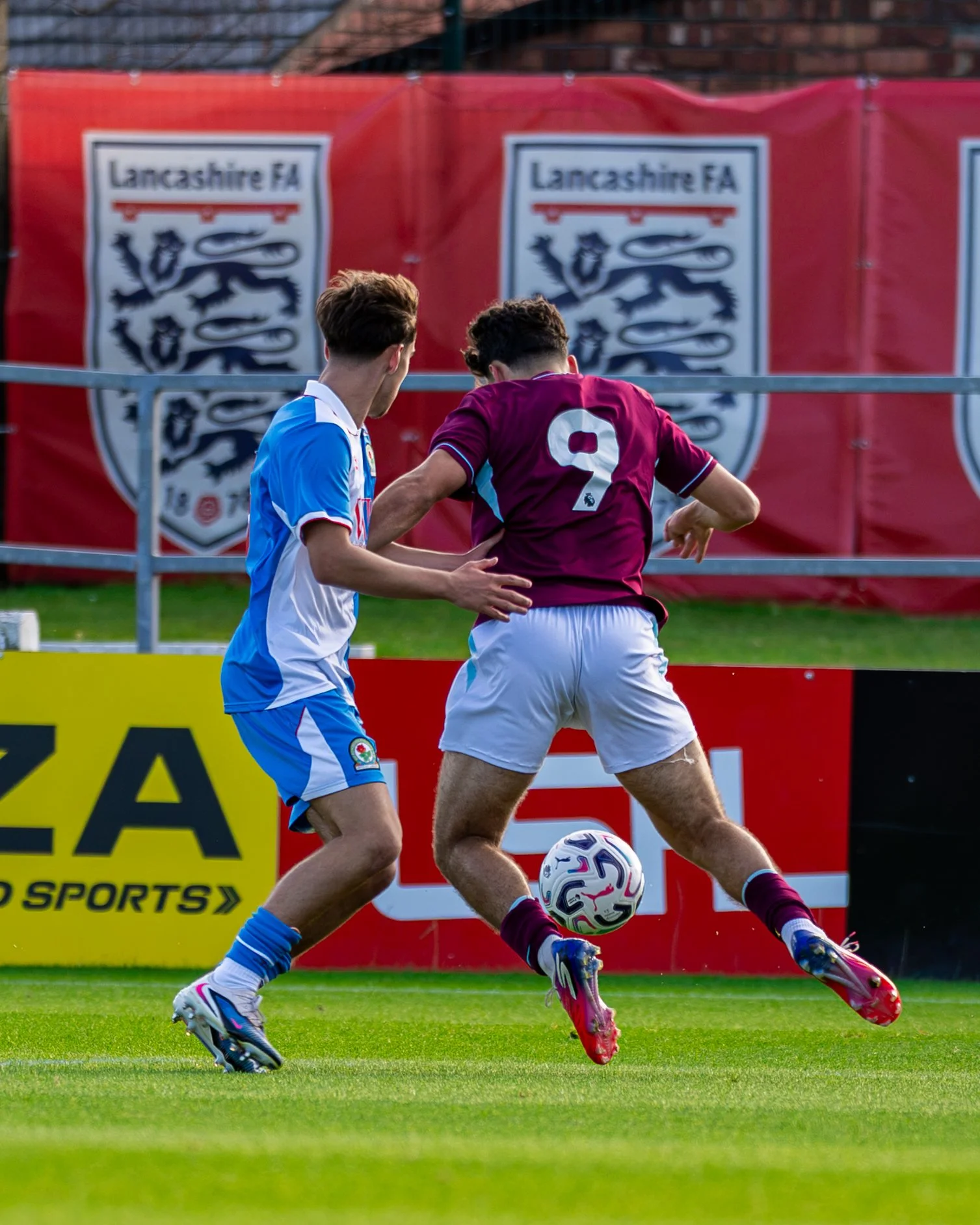 Two soccer players competing for the ball on a green field, with a large Lancashire FA banner in the background.