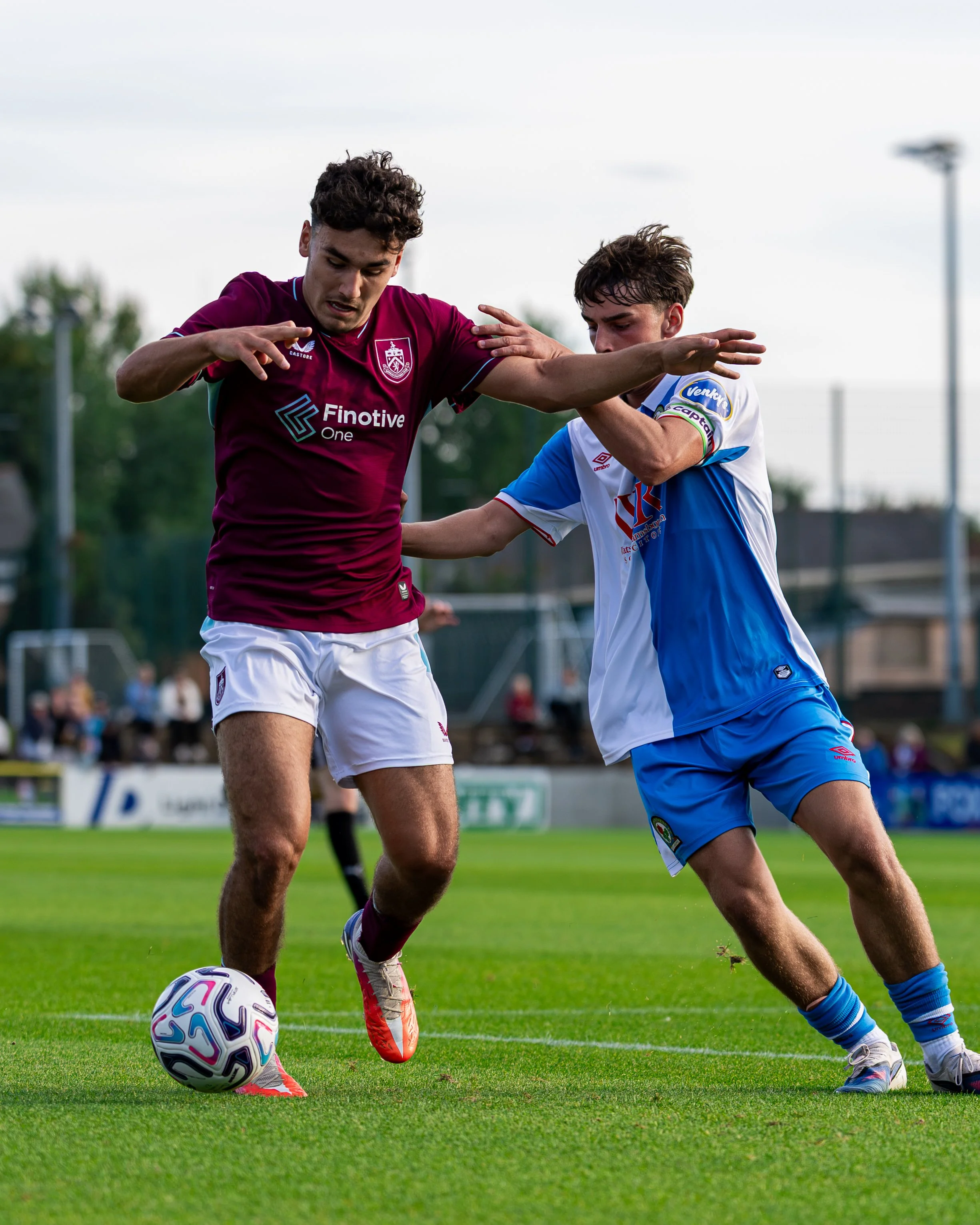 Two soccer players, one in a maroon jersey and the other in a blue and white jersey, compete for the ball on a grassy field during a match.