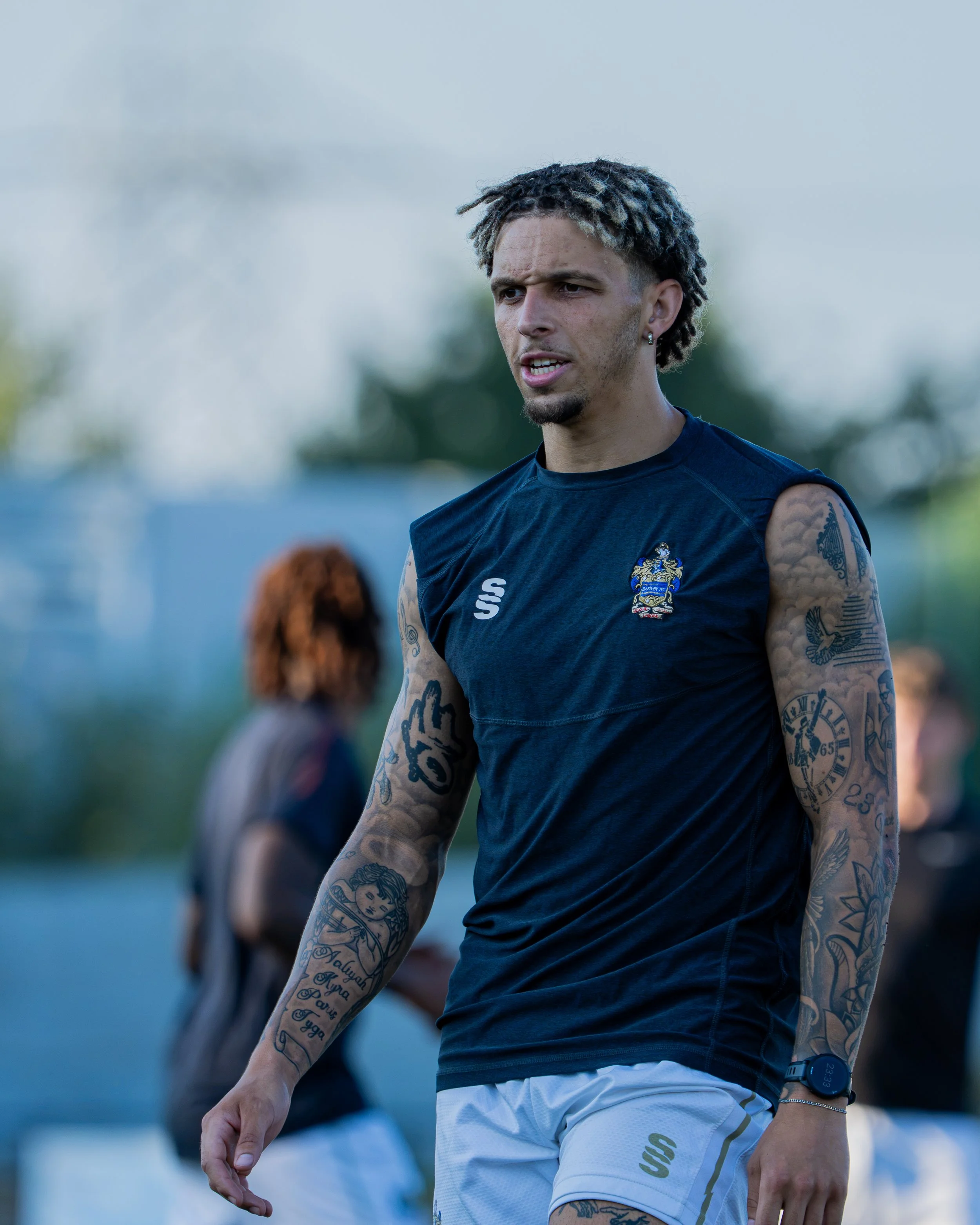 A male athlete with tattoos on his arms and shoulder, wearing a navy blue sleeveless shirt with a logo, white shorts, and a smartwatch, outdoors on a sunny day.