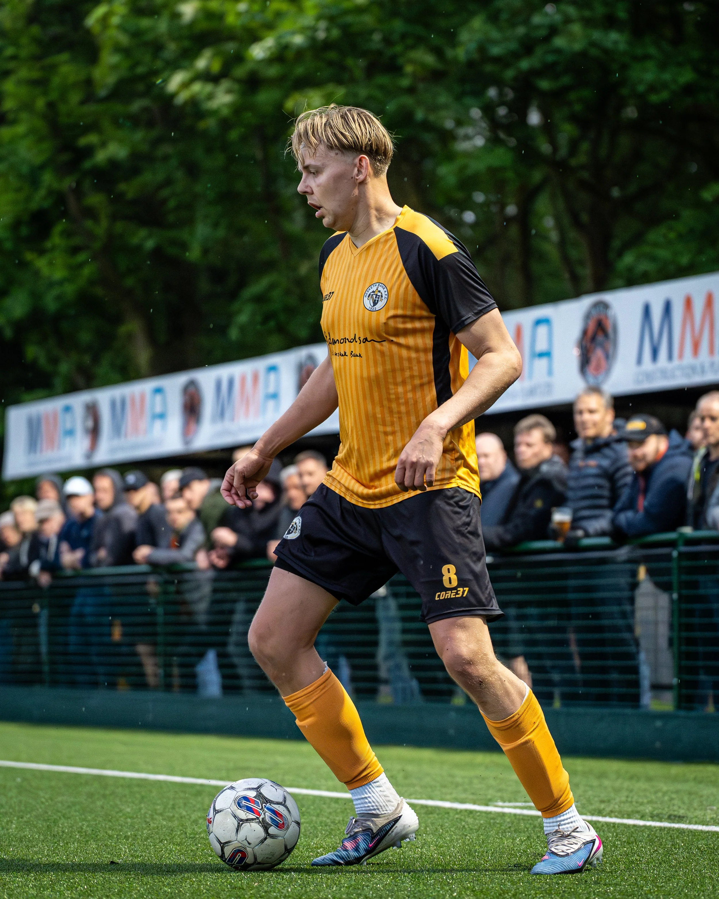 A young male soccer player in a yellow and black jersey with the number 8 and the logo 'CORE37' on it, about to kick a soccer ball on a green field with spectators and trees in the background.