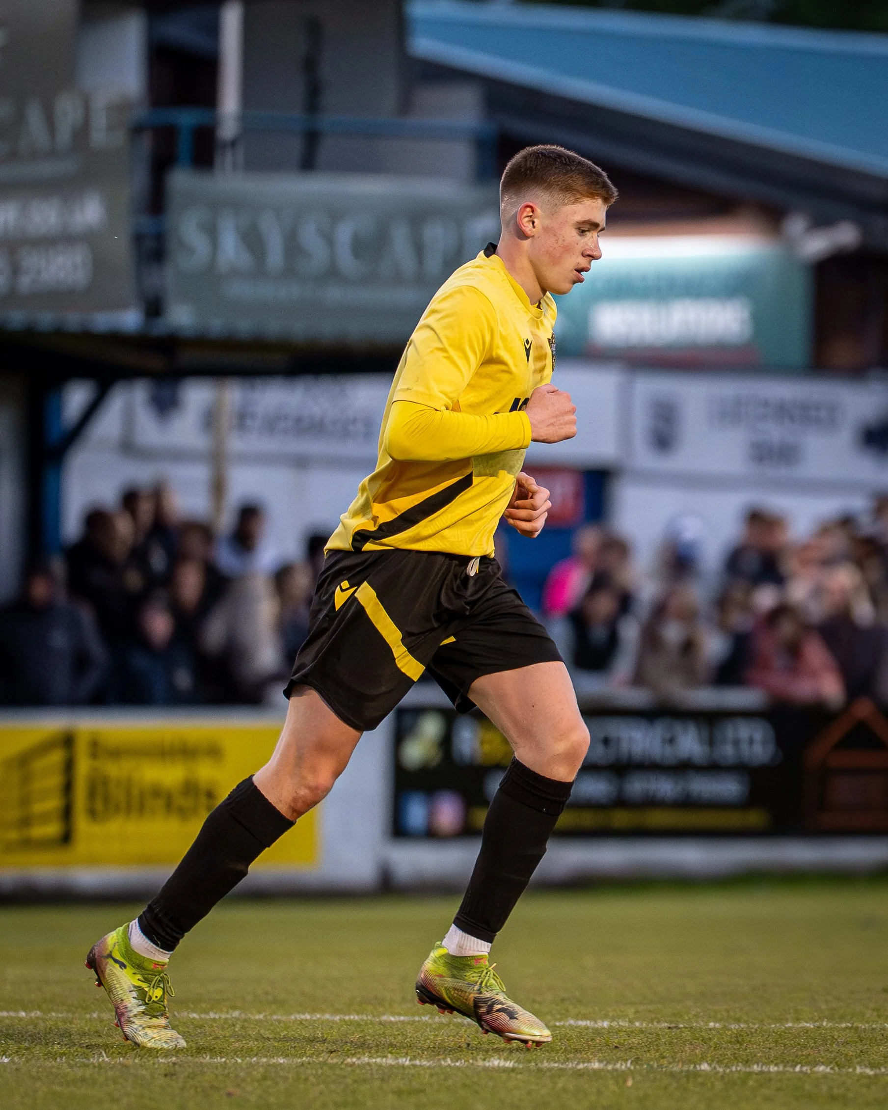 A young male soccer player in a yellow jersey, black shorts, and yellow cleats runs on the field during a game, with a focused expression and a crowd watching in the background.