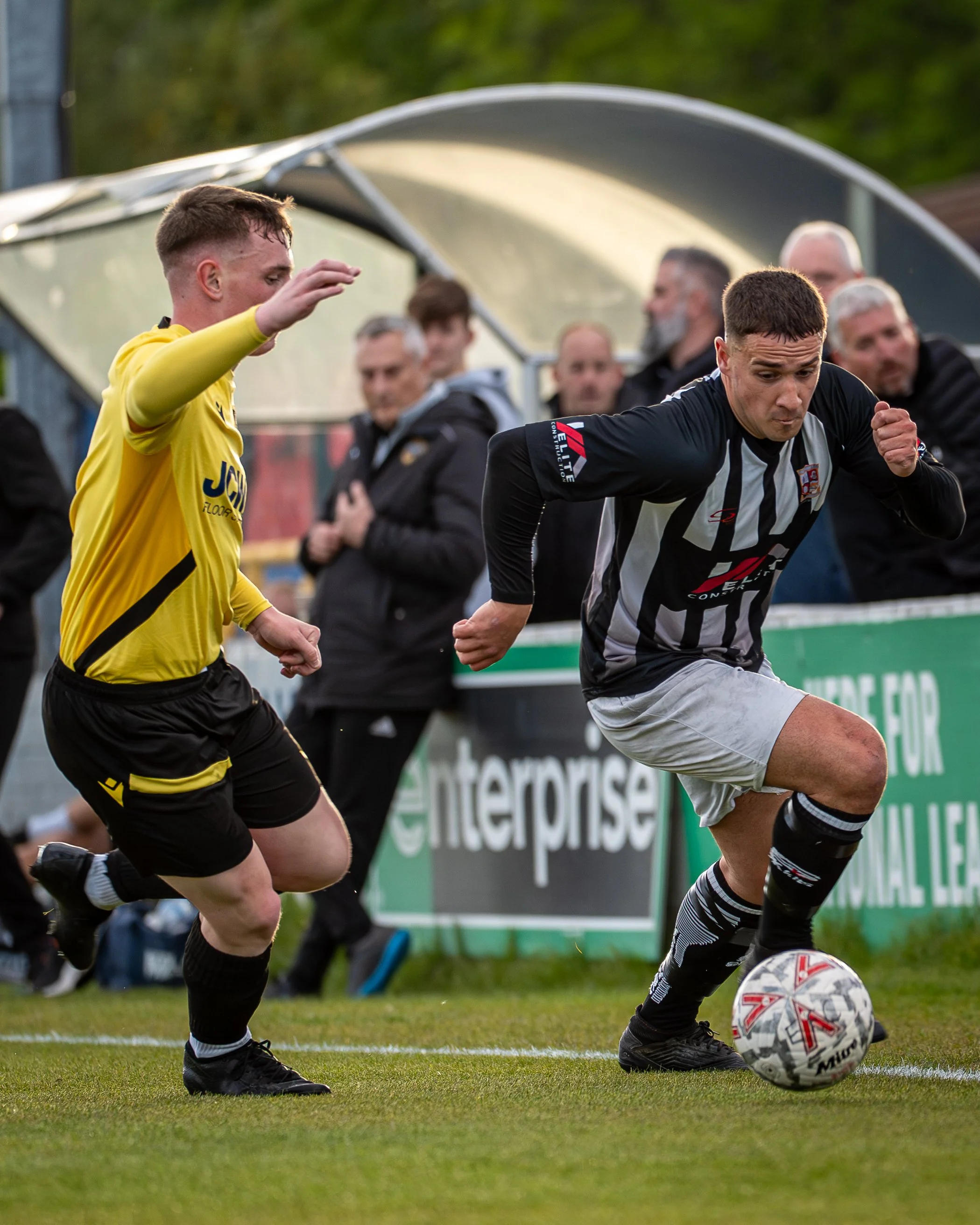 Two soccer players compete for the ball on the field during a match, with spectators and a dugout in the background.