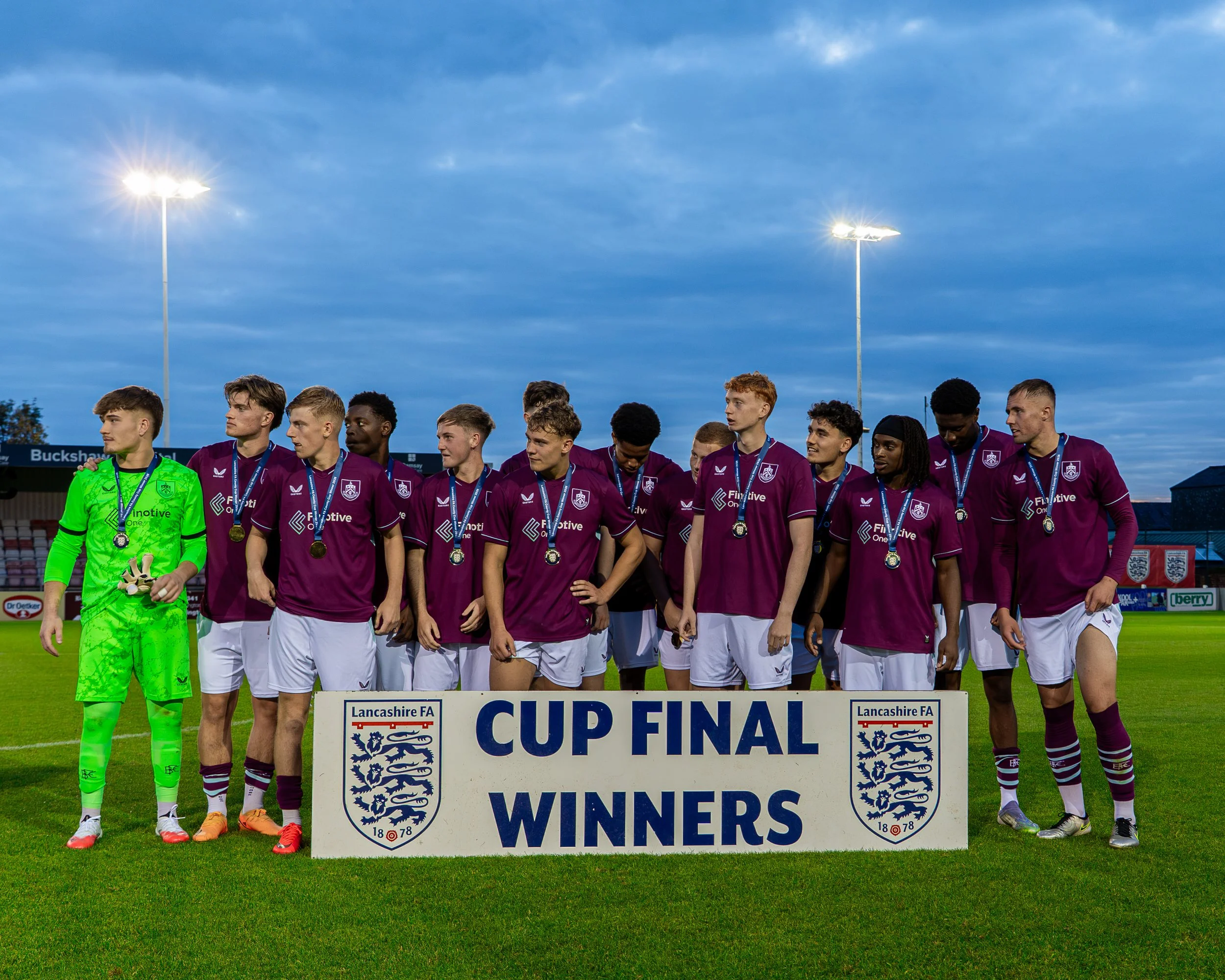 A group of soccer players celebrating on the field under cloudy skies after winning a cup final, standing behind a sign that reads 'Cup Final Winners'.