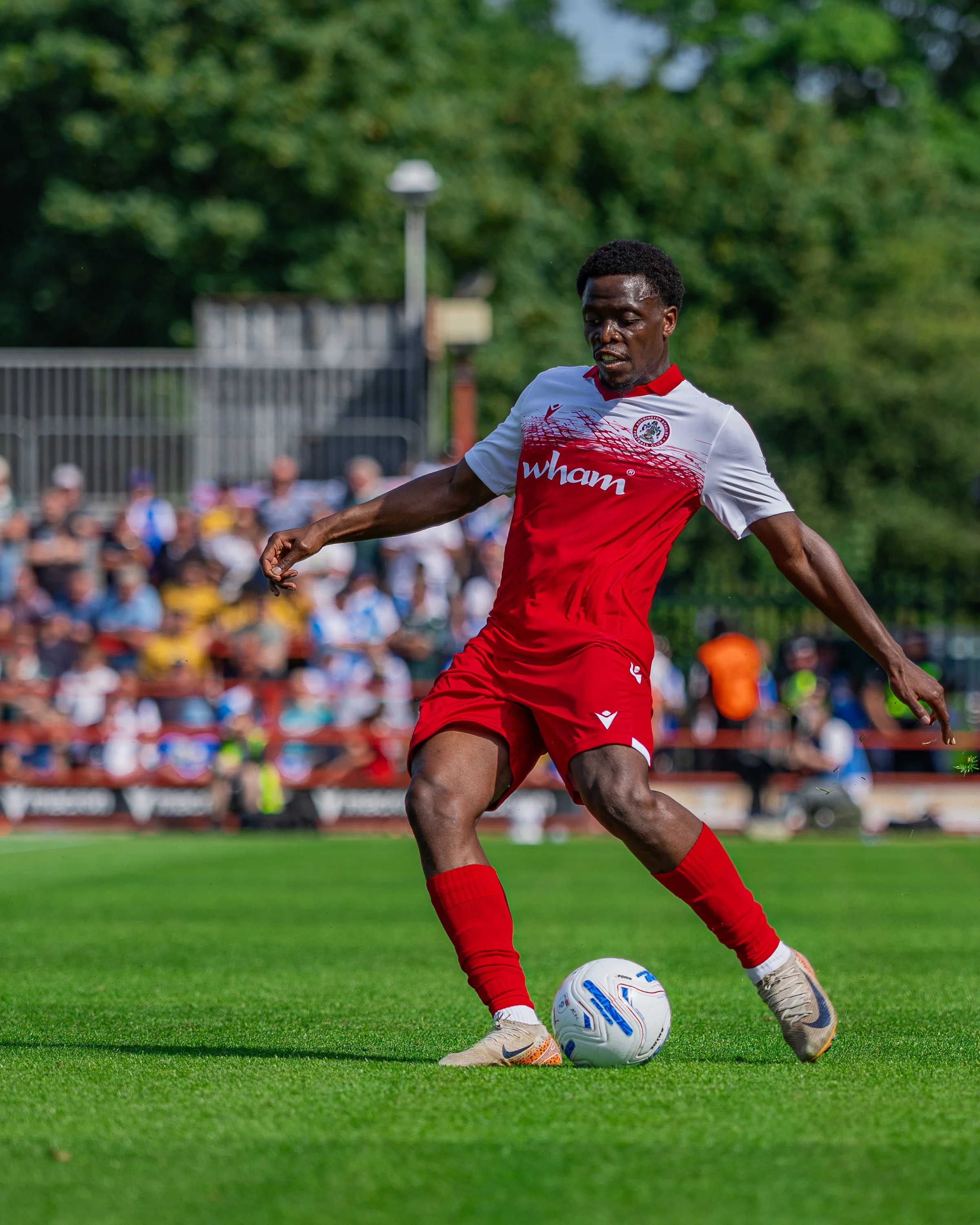 A soccer player in a red and white jersey is kicking a soccer ball on a green field during a game. The player is wearing beige Nike cleats and red socks, with a crowd of spectators blurred in the background.