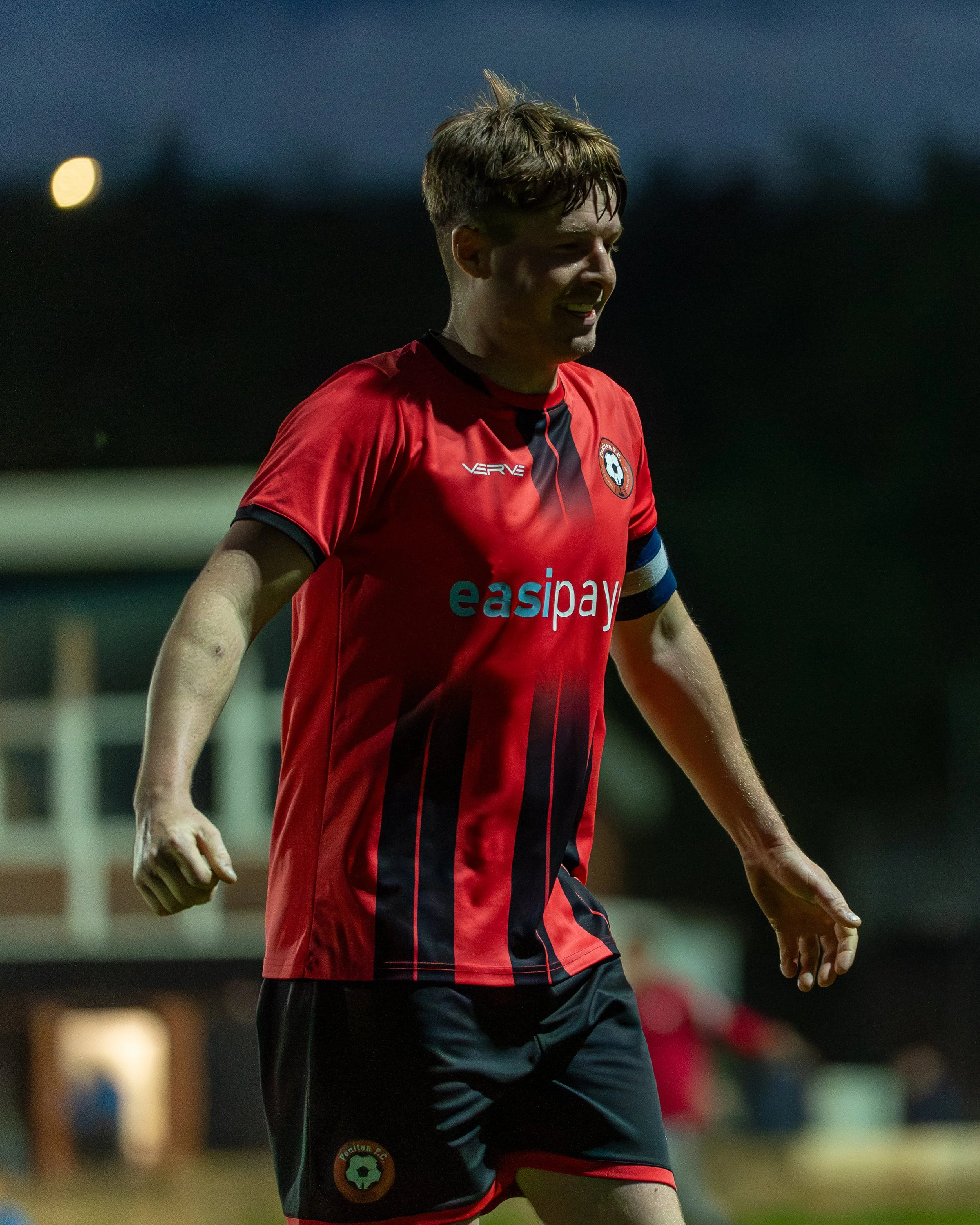 A male soccer player in a red and black uniform smiling, during a game at night.