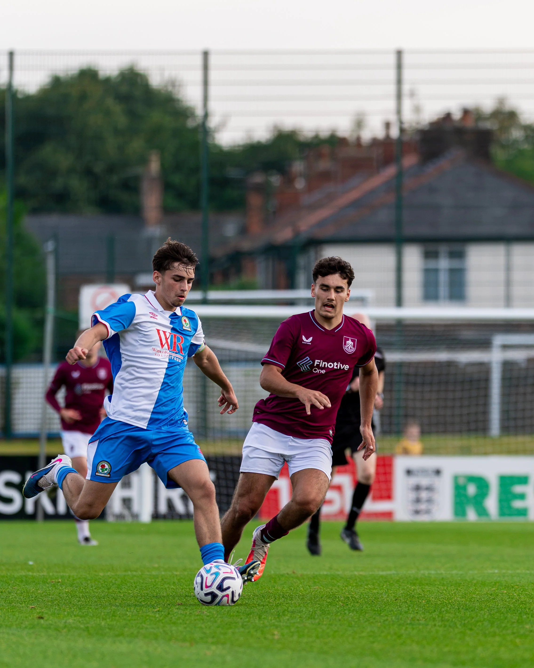 Two soccer players running on a green field, one in a blue and white uniform and the other in a maroon uniform, competing for the ball during a game, with a chain-link fence and houses visible in the background.