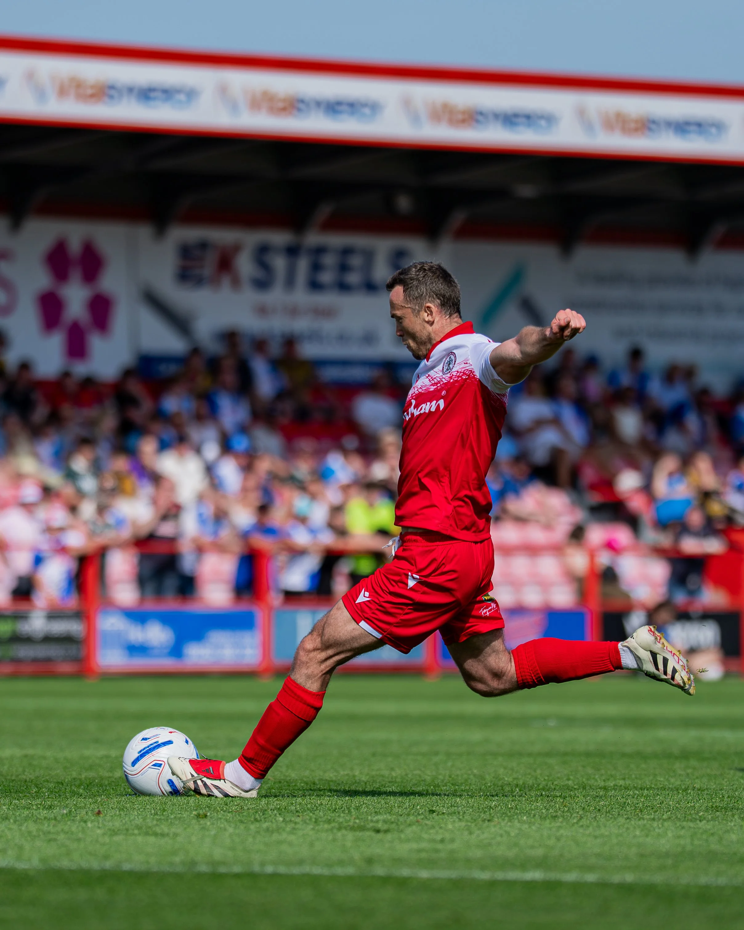 A soccer player in a red and white uniform kicking a soccer ball on a green grass field during a game with spectators in the background.