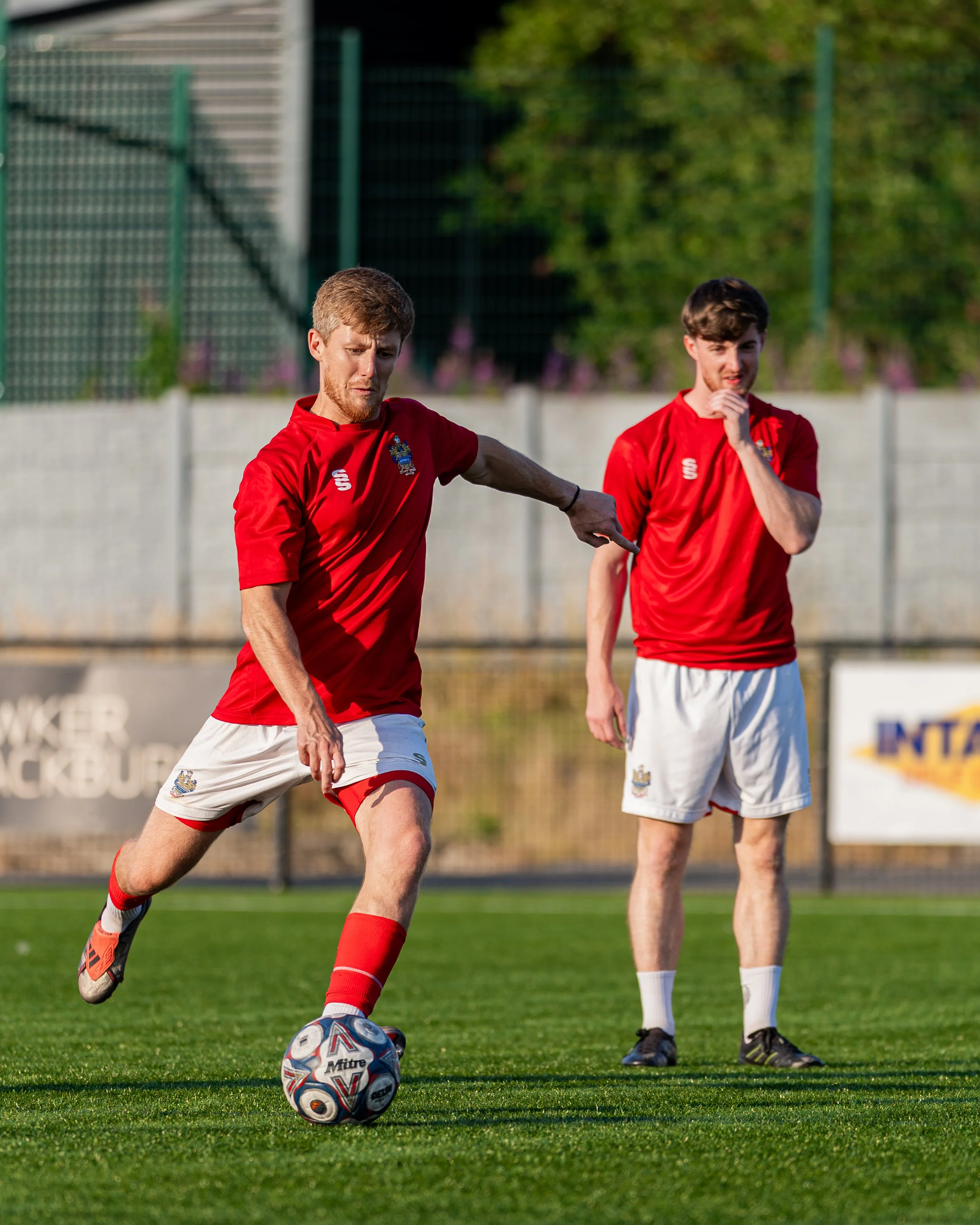Two young men in red athletic jerseys playing soccer on a grass field during daytime. One is kicking the ball, and the other is watching.