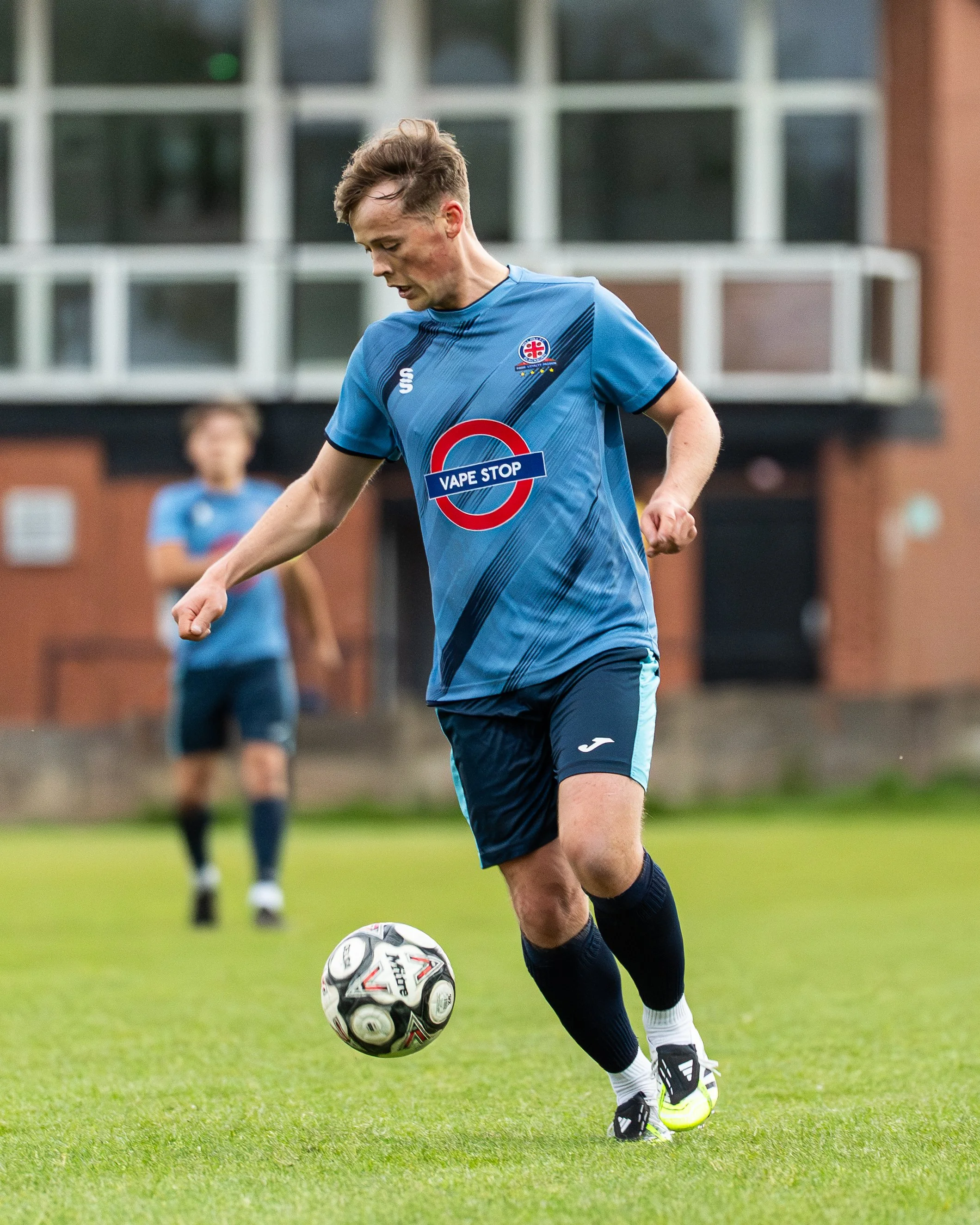A young male soccer player in a blue jersey with the logo 'Vape Stop' on the front, black shorts, and neon yellow and black cleats is on a grassy field, controlling a white and black soccer ball with his right foot. There is a blurred player in the b