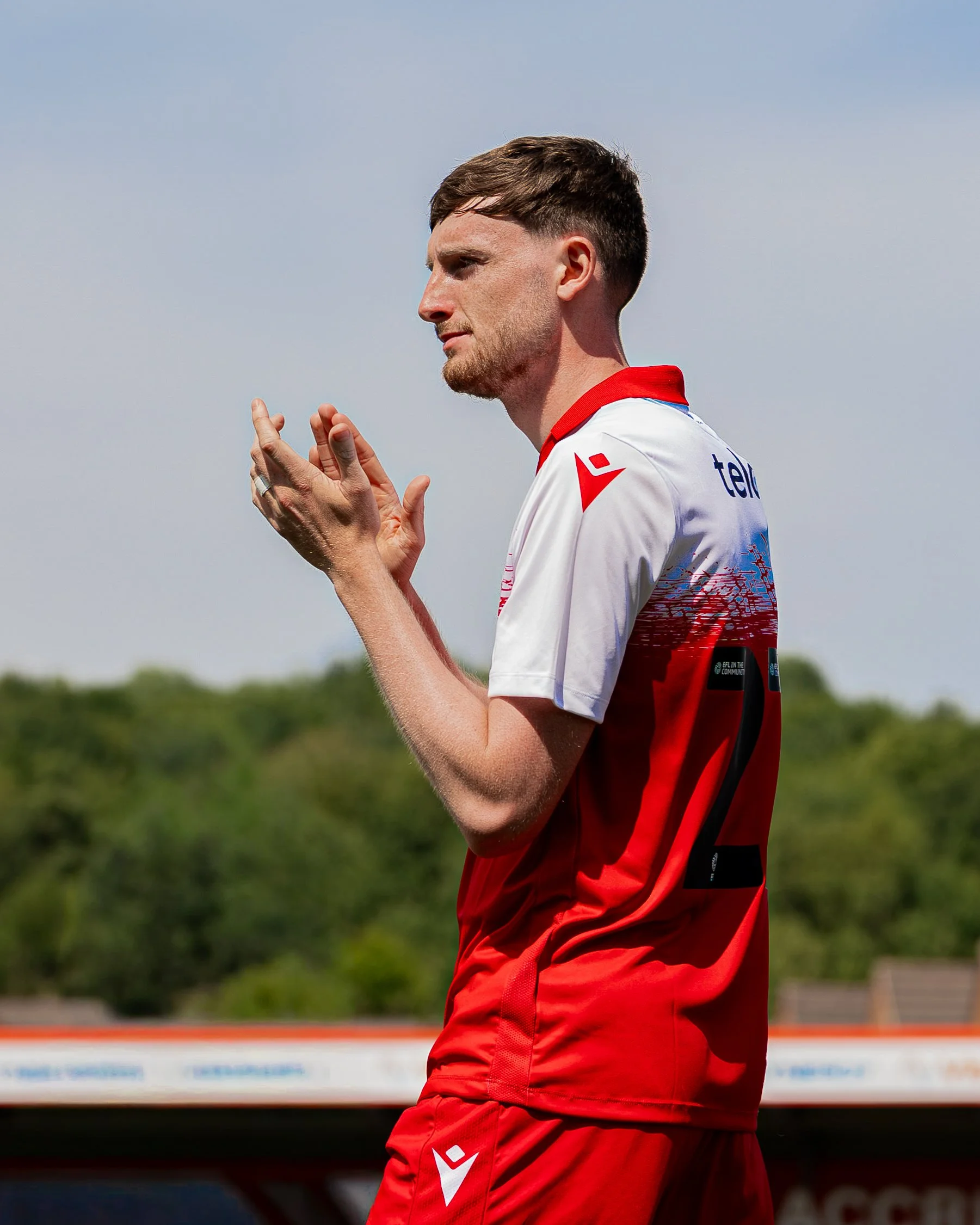 A man with short brown hair, facial hair, and light skin wearing a red and white sports jersey and shorts, standing outdoors against a background of blue sky and green trees, with hands clasped together.