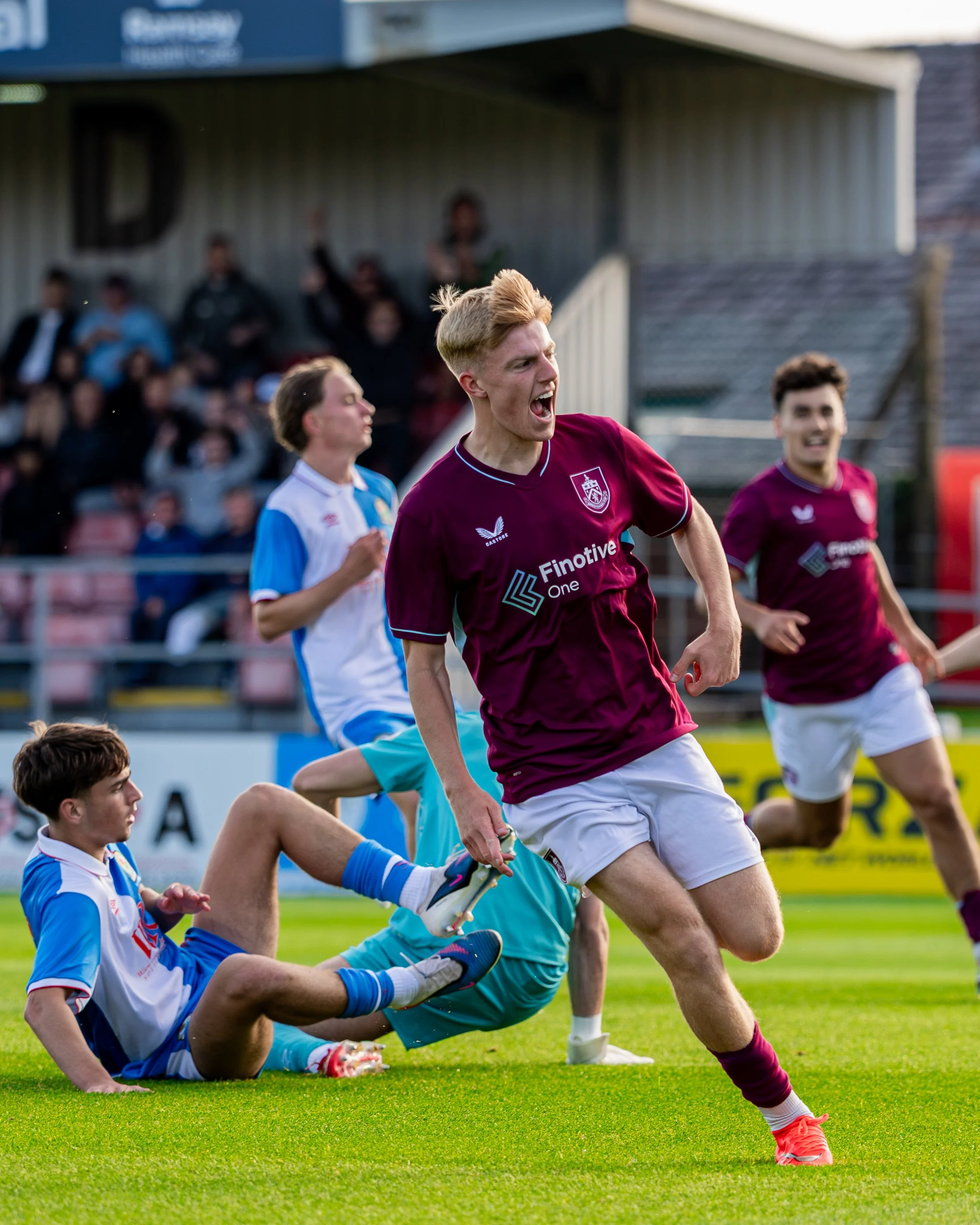 A soccer match with players on the field, focusing on a player in a maroon jersey celebrating a goal, surrounded by players in blue and white jerseys, with spectators in the background.