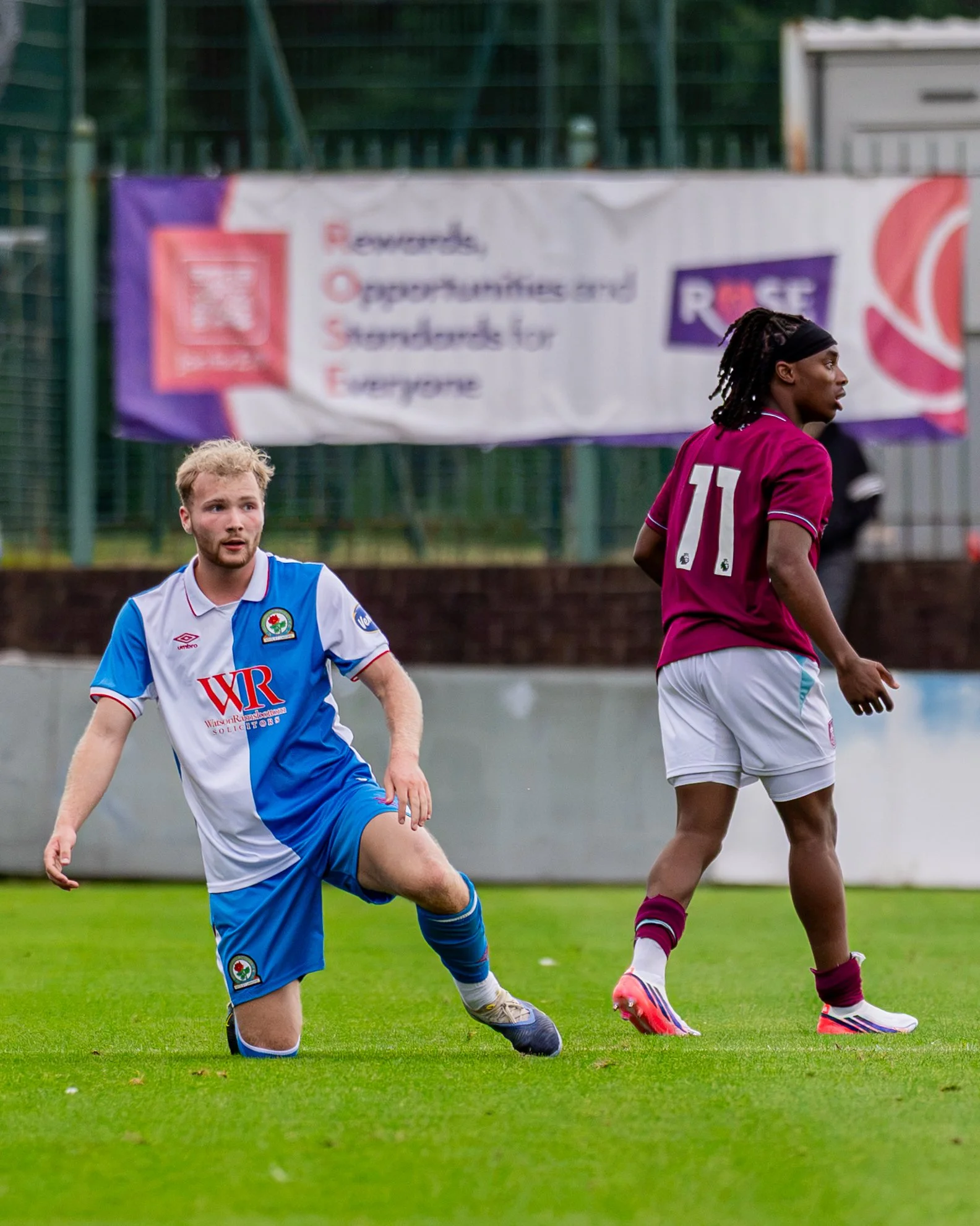 Two soccer players, one in a white and blue uniform and the other in a maroon uniform, playing on a grass field with a fence and a banner in the background.