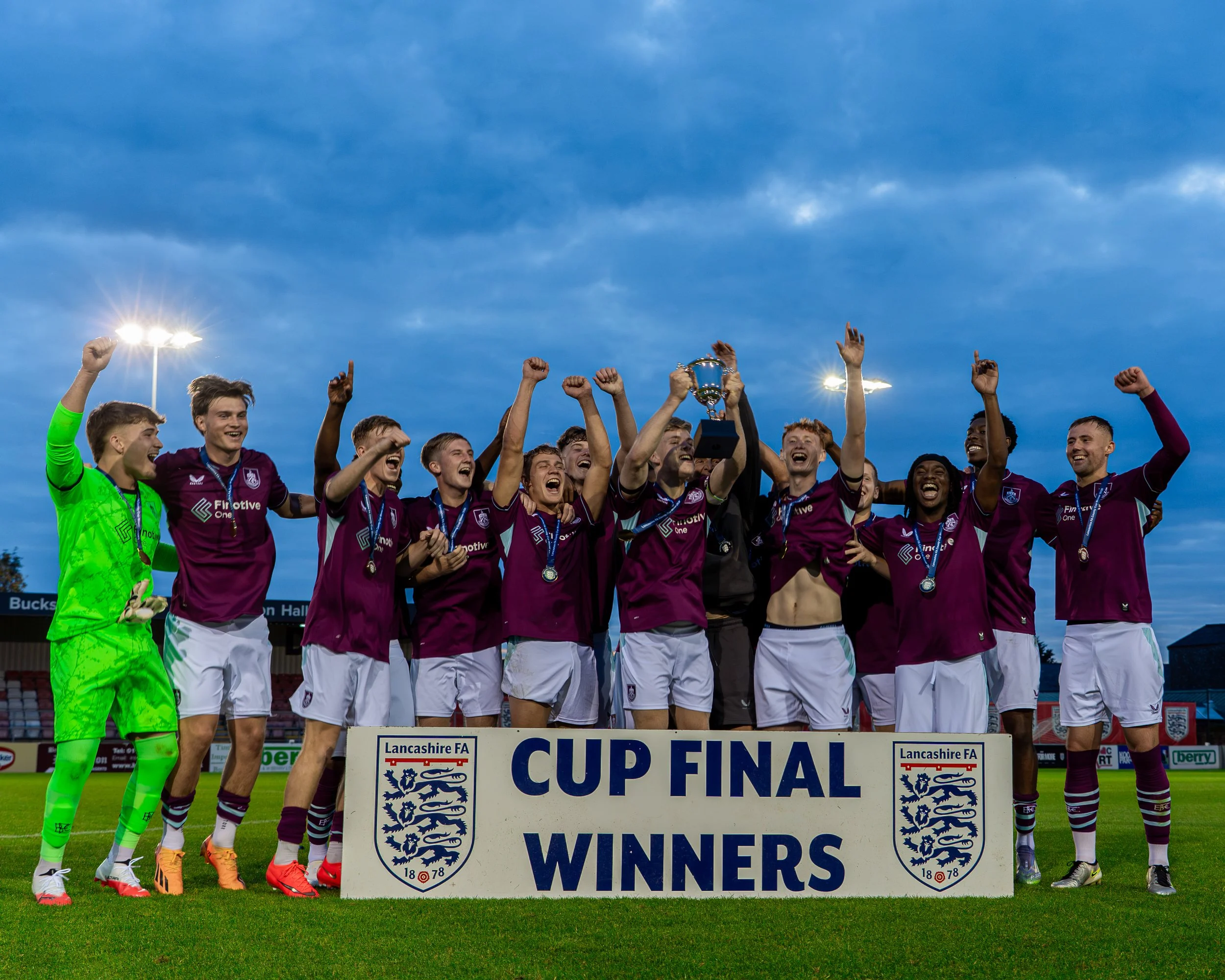 A group of soccer players celebrating a victory on the field, holding a trophy, standing behind a sign that reads 'Cup Final Winners,' with a blue sky and stadium lights overhead.