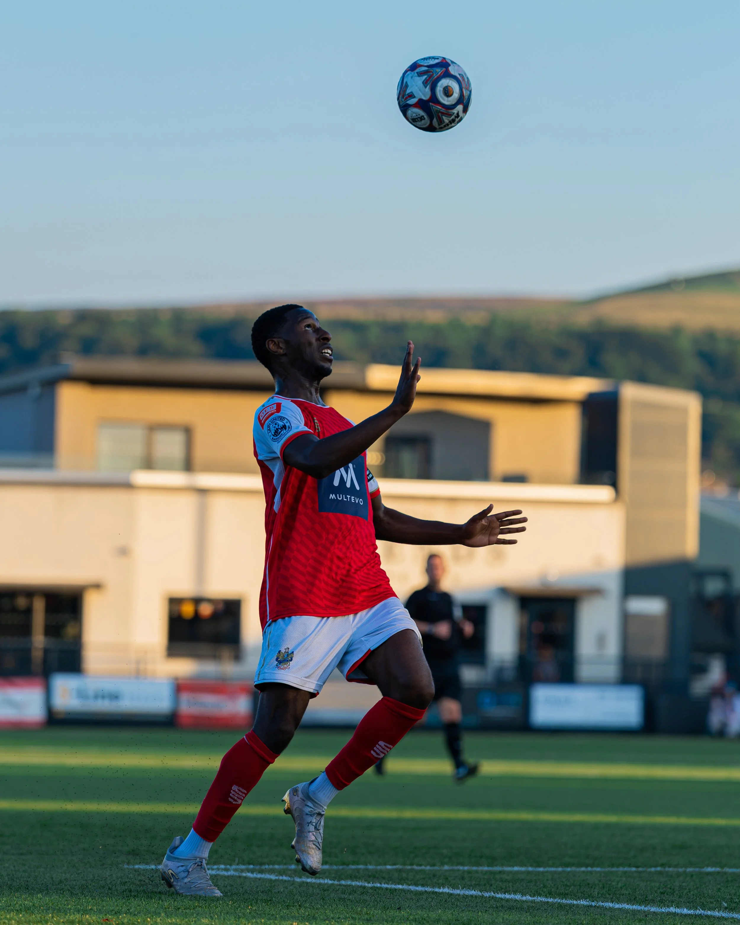 A soccer player wearing a red and white uniform is preparing to kick a colorful soccer ball in mid-air during a match on a green field. The background shows a building and distant hills in the late afternoon sunlight.