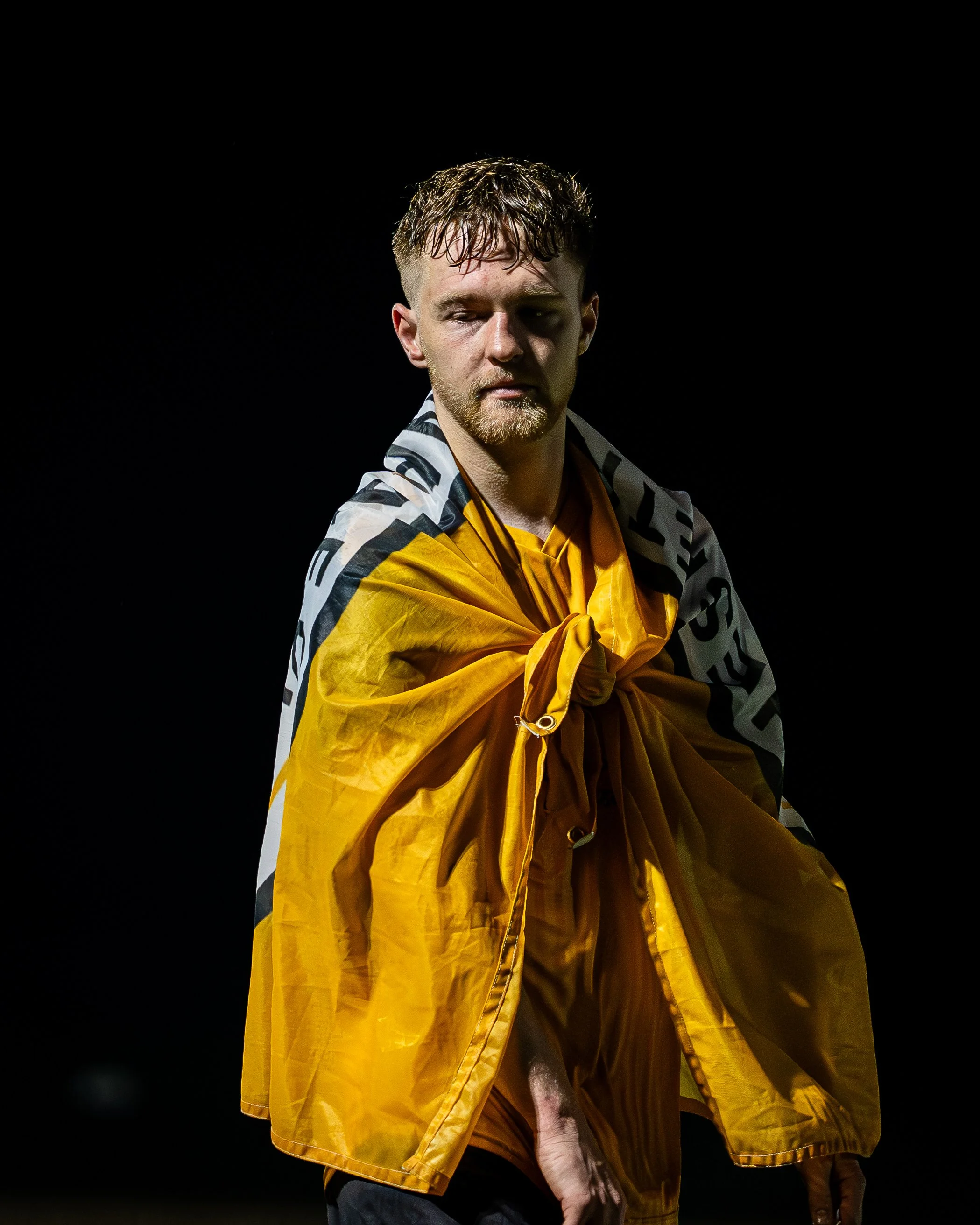 A young man with wet hair and a beard, wearing a yellow shirt and a black-and-white patterned jacket, looking down with a serious expression, against a black background.