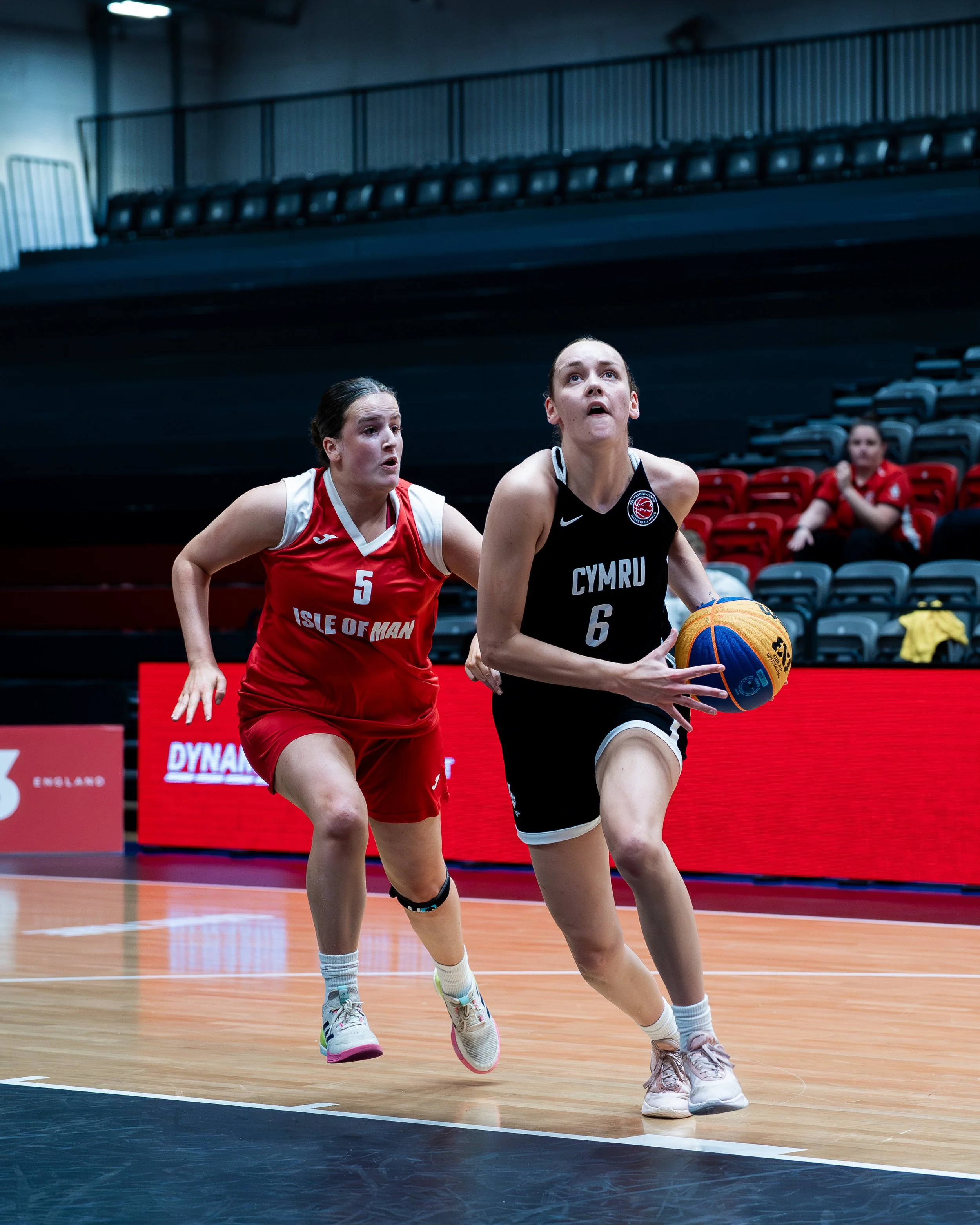 Two female basketball players, one in a red uniform representing Isle of Man and the other in black representing Wales, compete for the ball on an indoor court.
