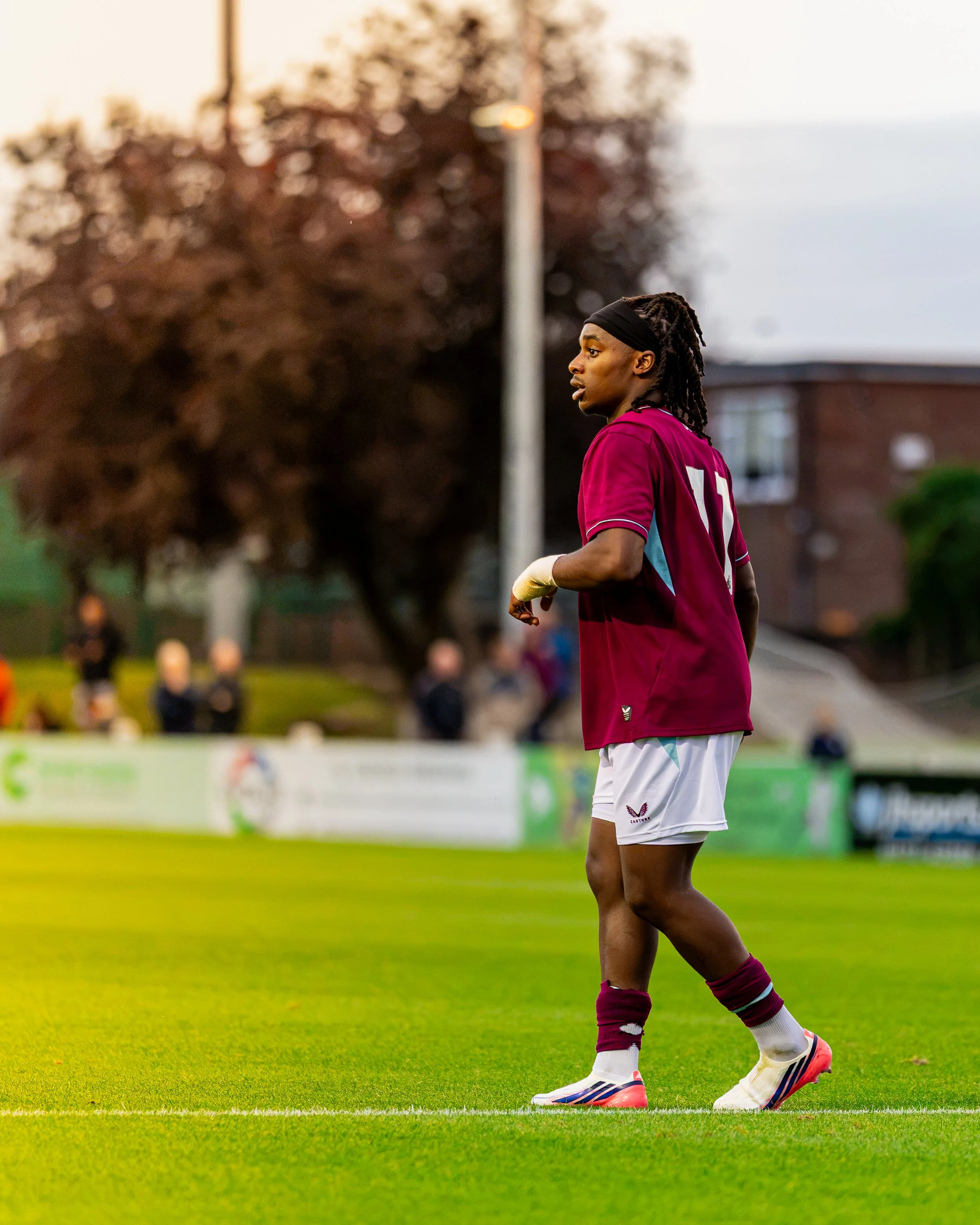 A soccer player in a maroon jersey and white shorts standing on a grass field, with spectators and trees in the background.