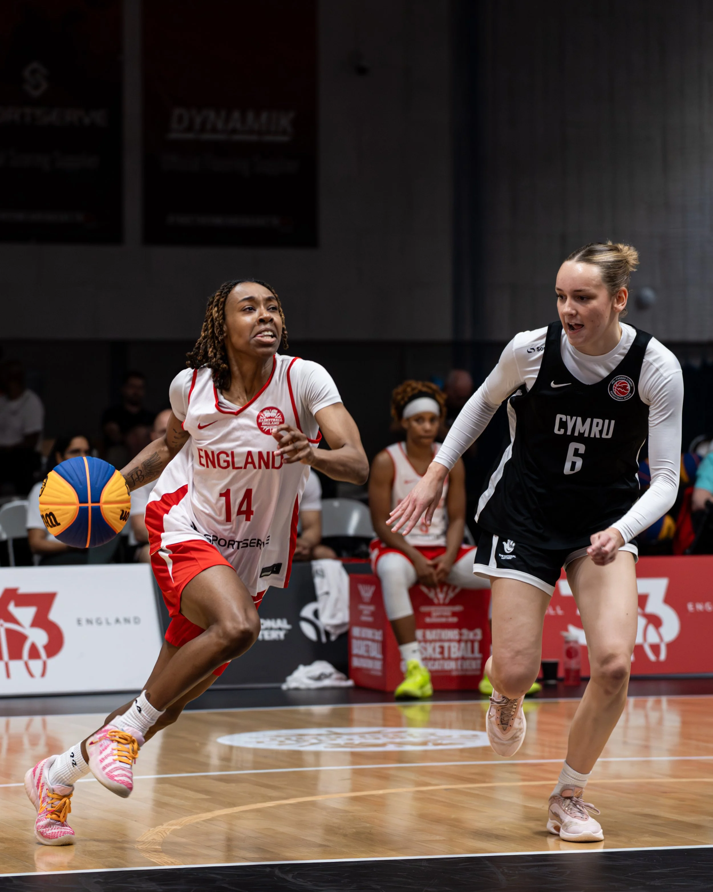 Two female basketball players competing on the court during a game, with one dribbling the ball and the other defending, in an indoor arena.