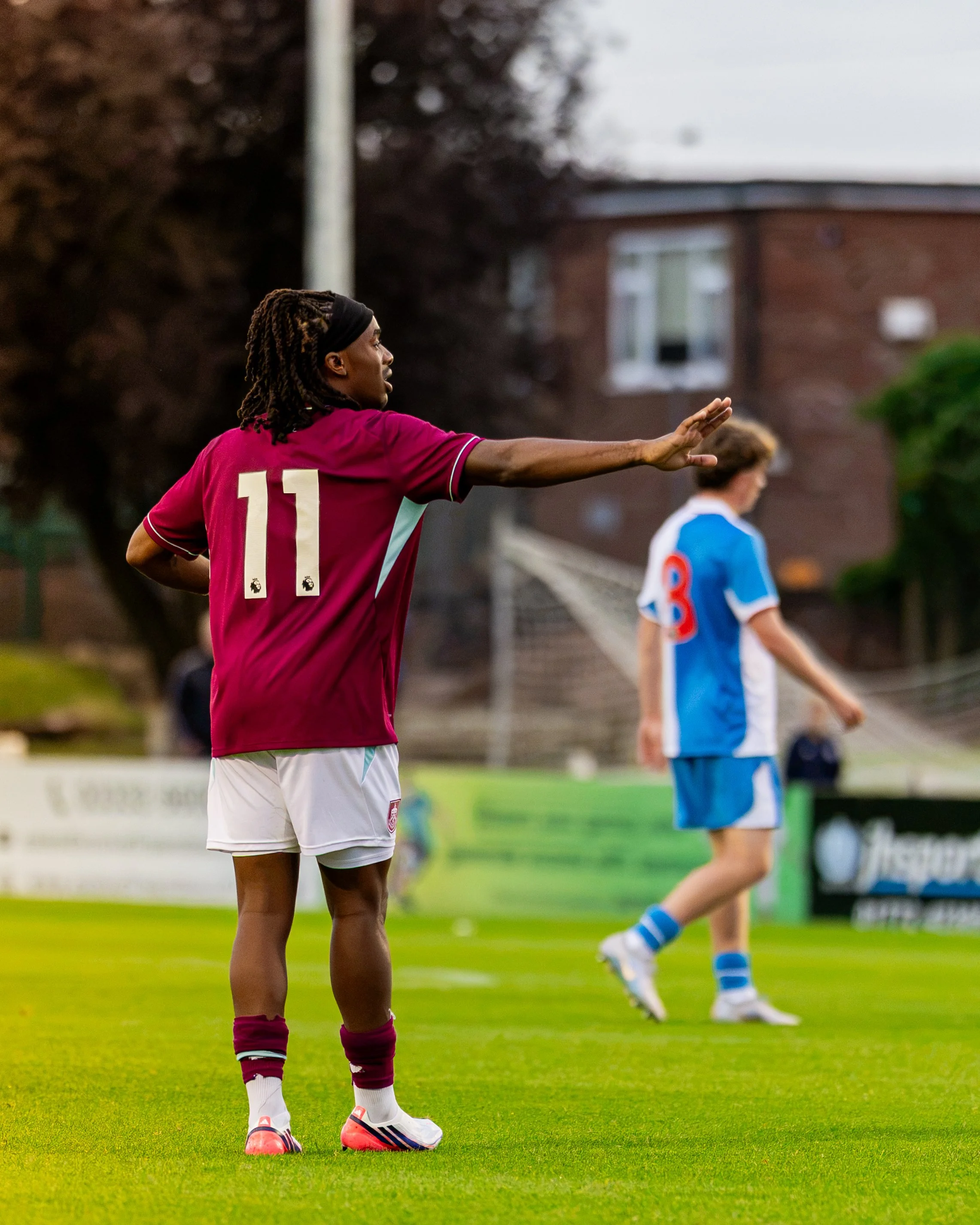 A female soccer player in a maroon jersey with the number 11, white shorts, and pink Nike soccer cleats, is standing on a green soccer field. She has long dreadlocks with a black headband and is extending her right arm with her hand raised. In the ba
