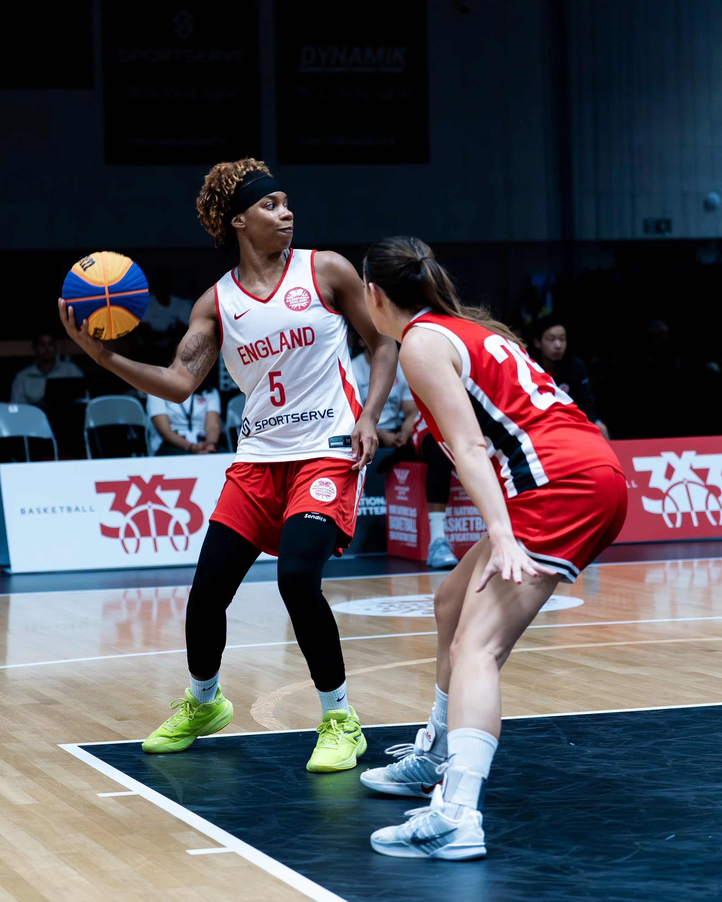 Two female basketball players from England team in red and white uniforms on indoor court. One has the ball and is wearing neon green shoes; the other is crouching and wearing light gray shoes. Spectators and banners in background.