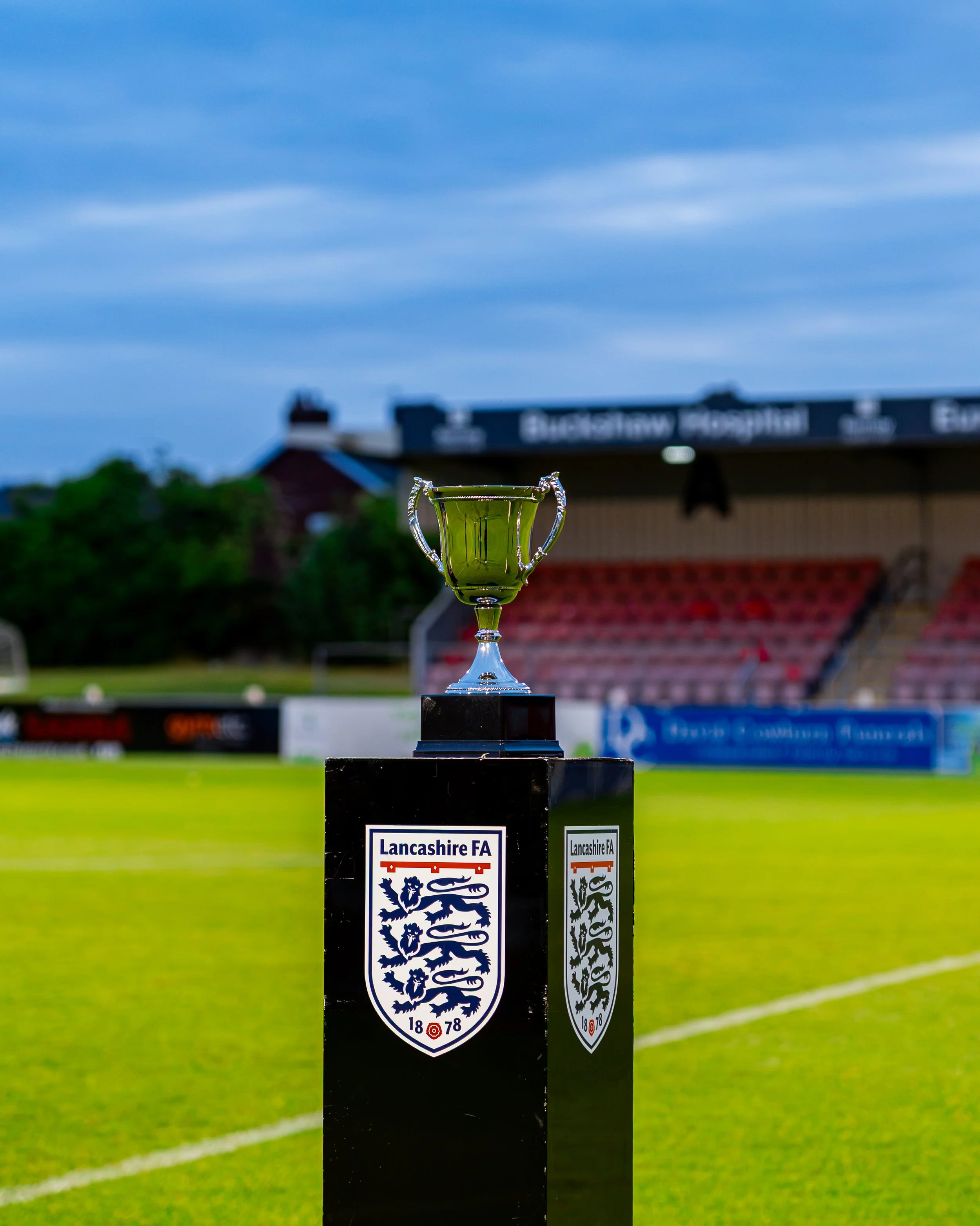 A trophy stands on a black pedestal on a sports field with a Lancaster FA logo, with a stadium and trees in the background under a partly cloudy sky.