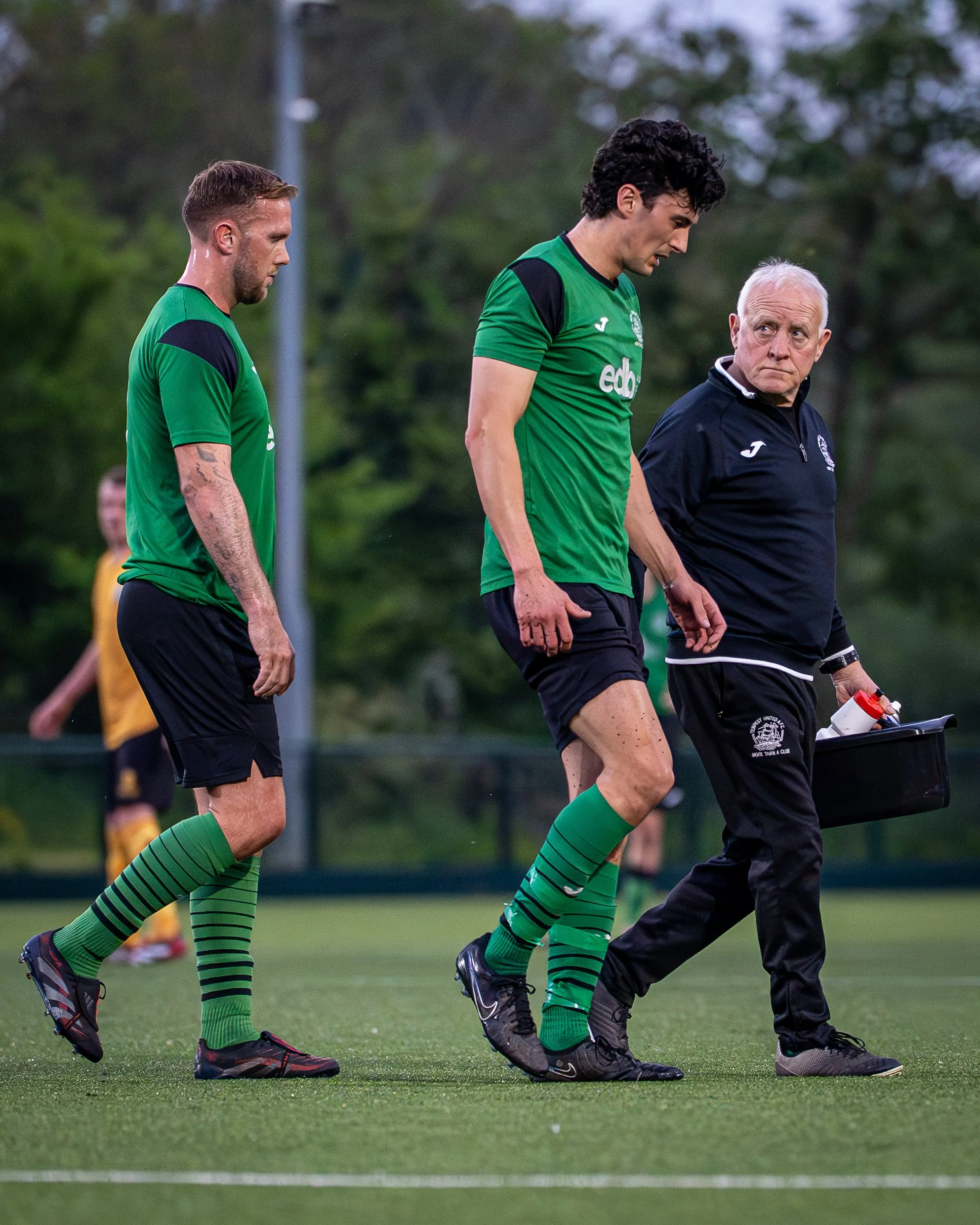 Three men walking on a sports field, two in green jerseys and one in a black jacket, with trees in the background.