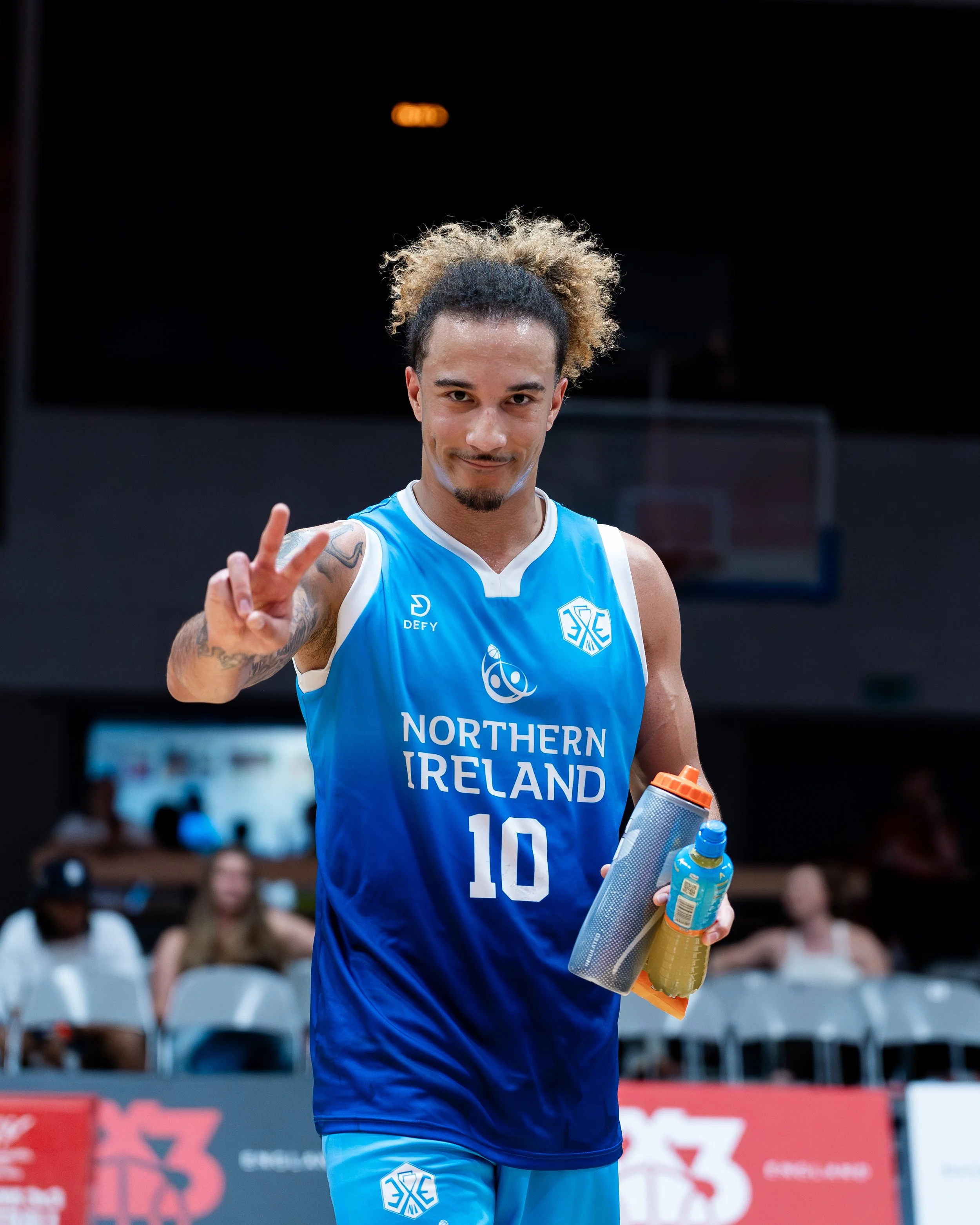 A male volleyball player in a blue jersey holding a water bottle and making a peace sign with his right hand, standing in an indoor sports arena.