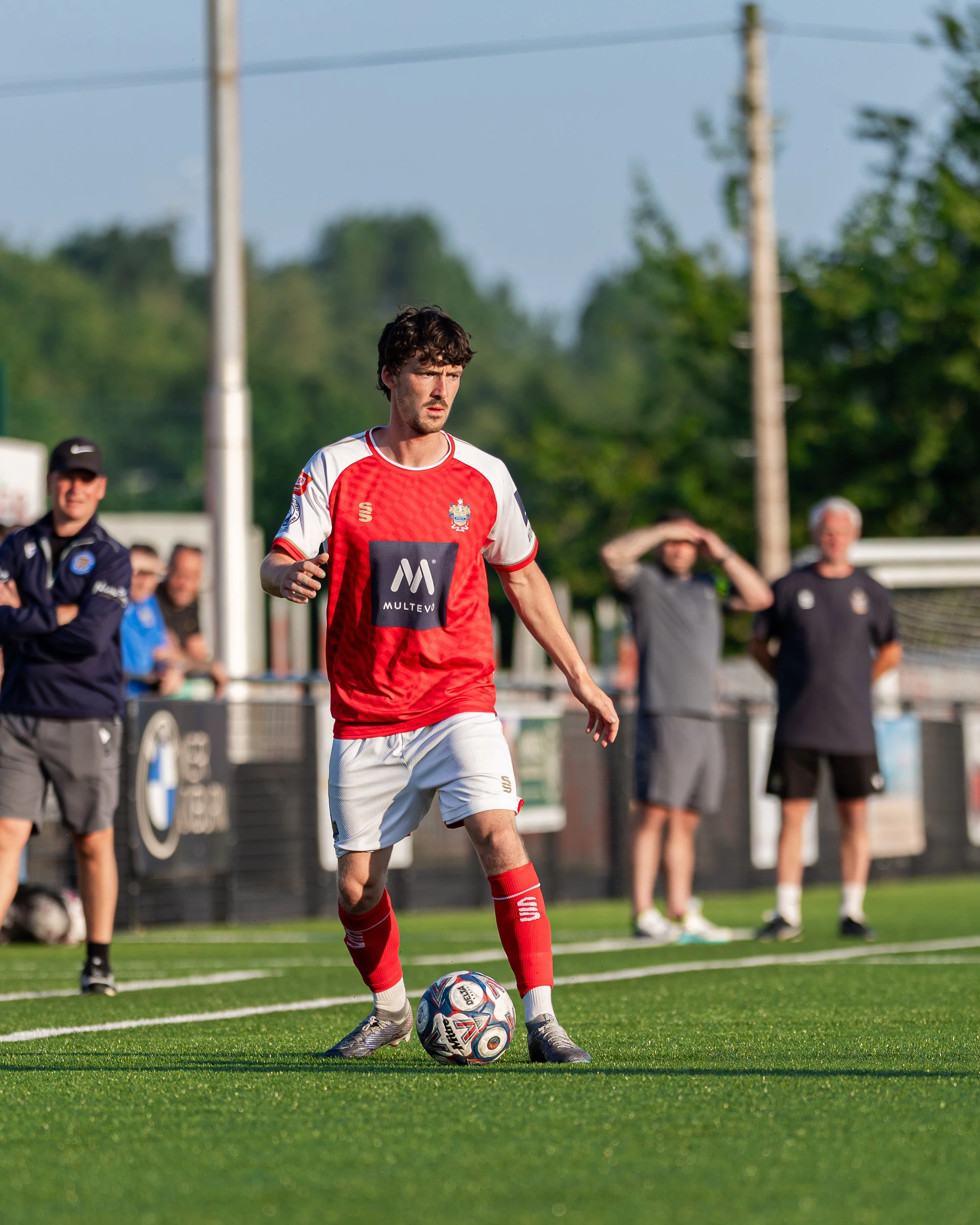 Young male soccer player in a red and white uniform preparing to kick a soccer ball on a green field during a game, while several spectators watch in the background.