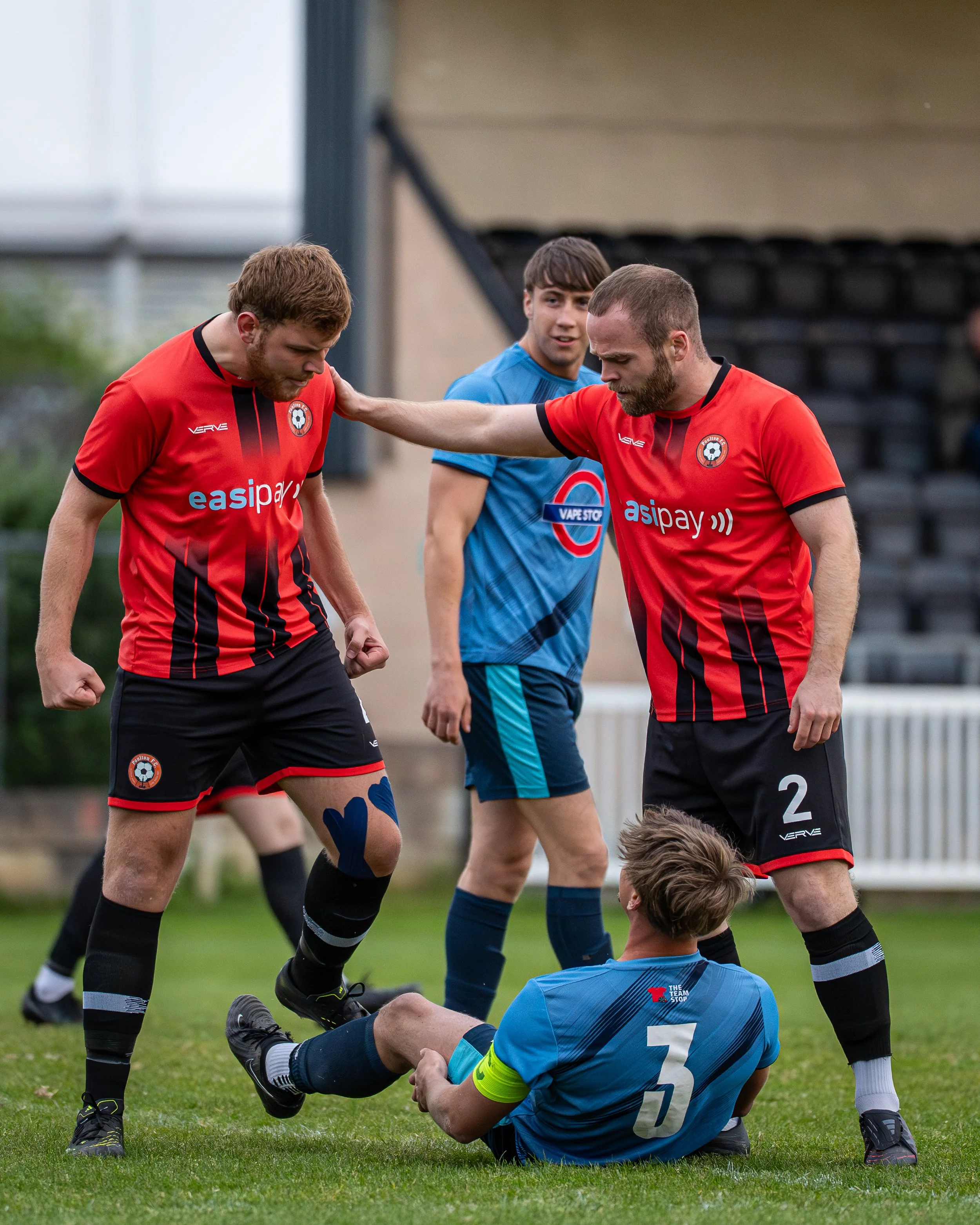 Two soccer players from opposing teams fight on the field, with one player sitting on the ground and holding his leg while another player helping him up. Two more players are in the background observing the scene.