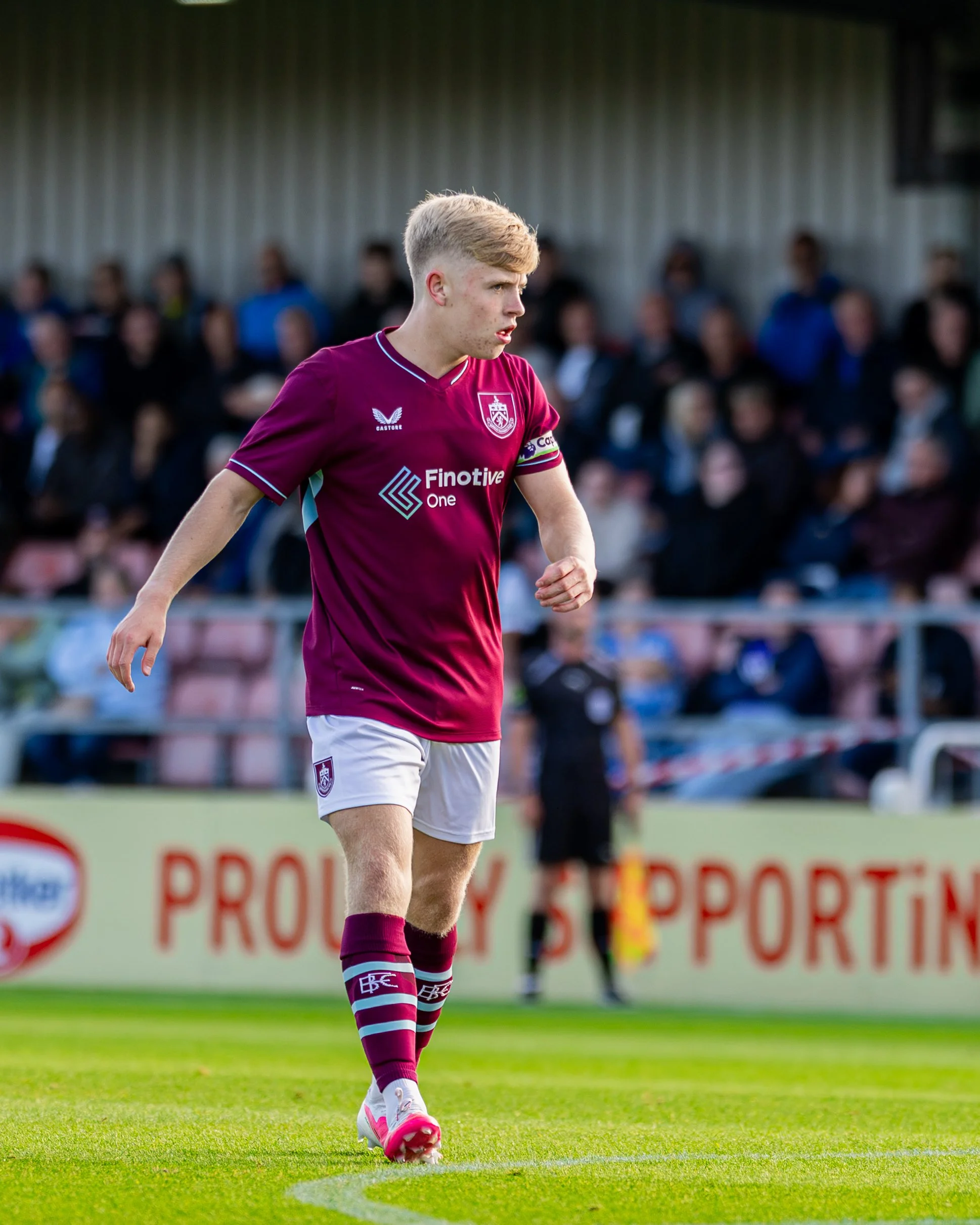 A young male soccer player with light blond hair in a fade haircut, wearing a maroon and white uniform with the logo of Brisbane Roar, is walking on the field during a game. In the background, there are spectators watching the match.