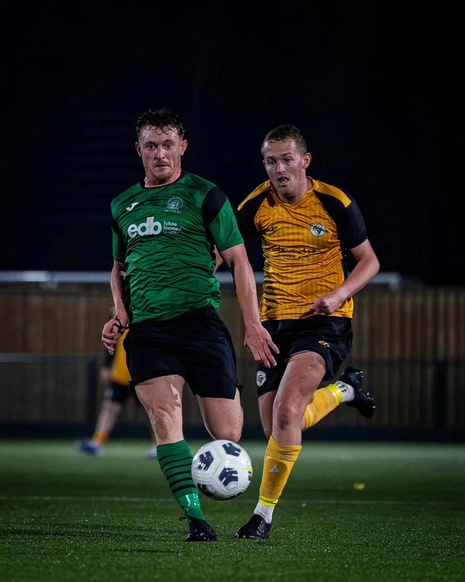 Two soccer players compete for the ball during a match, with one in a green jersey and the other in a yellow jersey, on a green turf field at night.