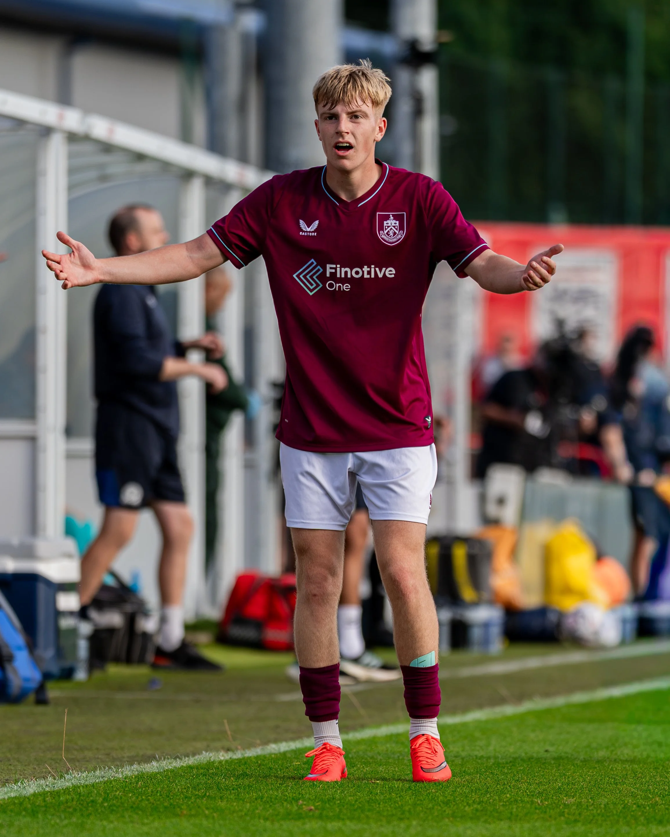 A young male soccer player wearing a maroon jersey, white shorts, maroon socks, and orange cleats stands on a soccer field with arms outstretched during a game.