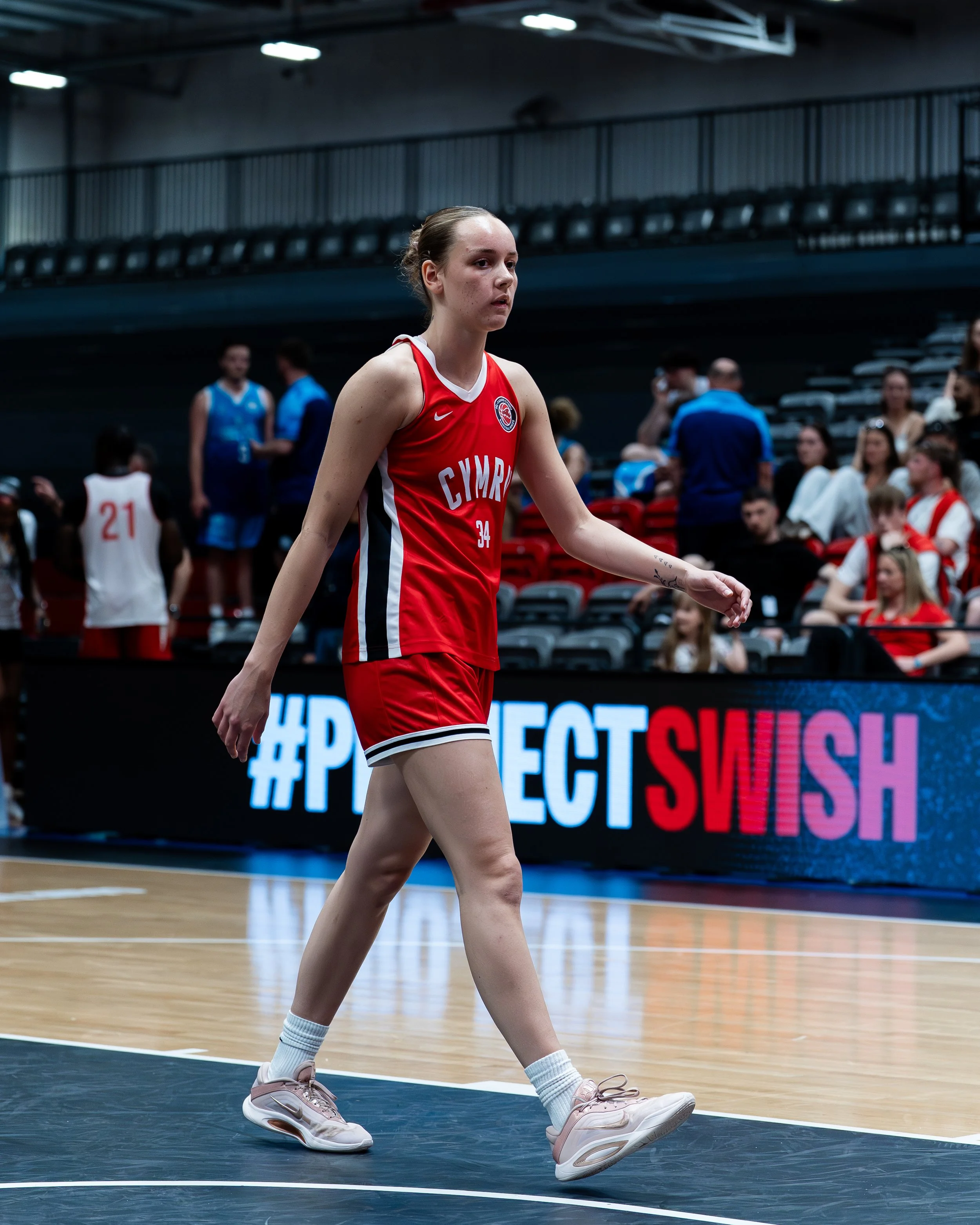 A female basketball player in a red uniform with the word 'Cymru' is walking on a basketball court during a game, with spectators and other players in the background.