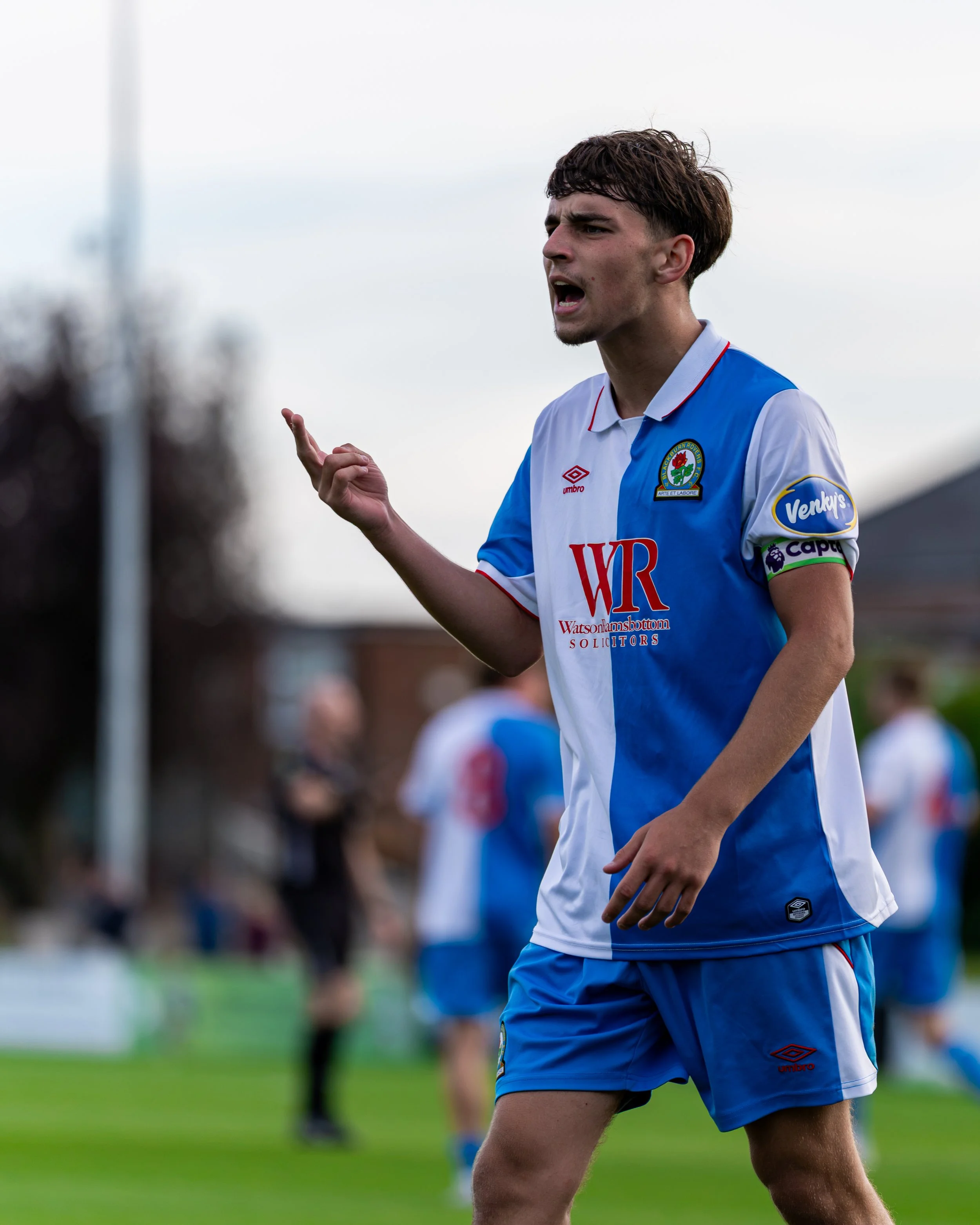 A young male soccer player with short, curly brown hair and light skin, wearing a blue and white jersey with various sponsor logos, is standing on the field during daytime gesturing with his right hand and speaking, with a slightly serious expression