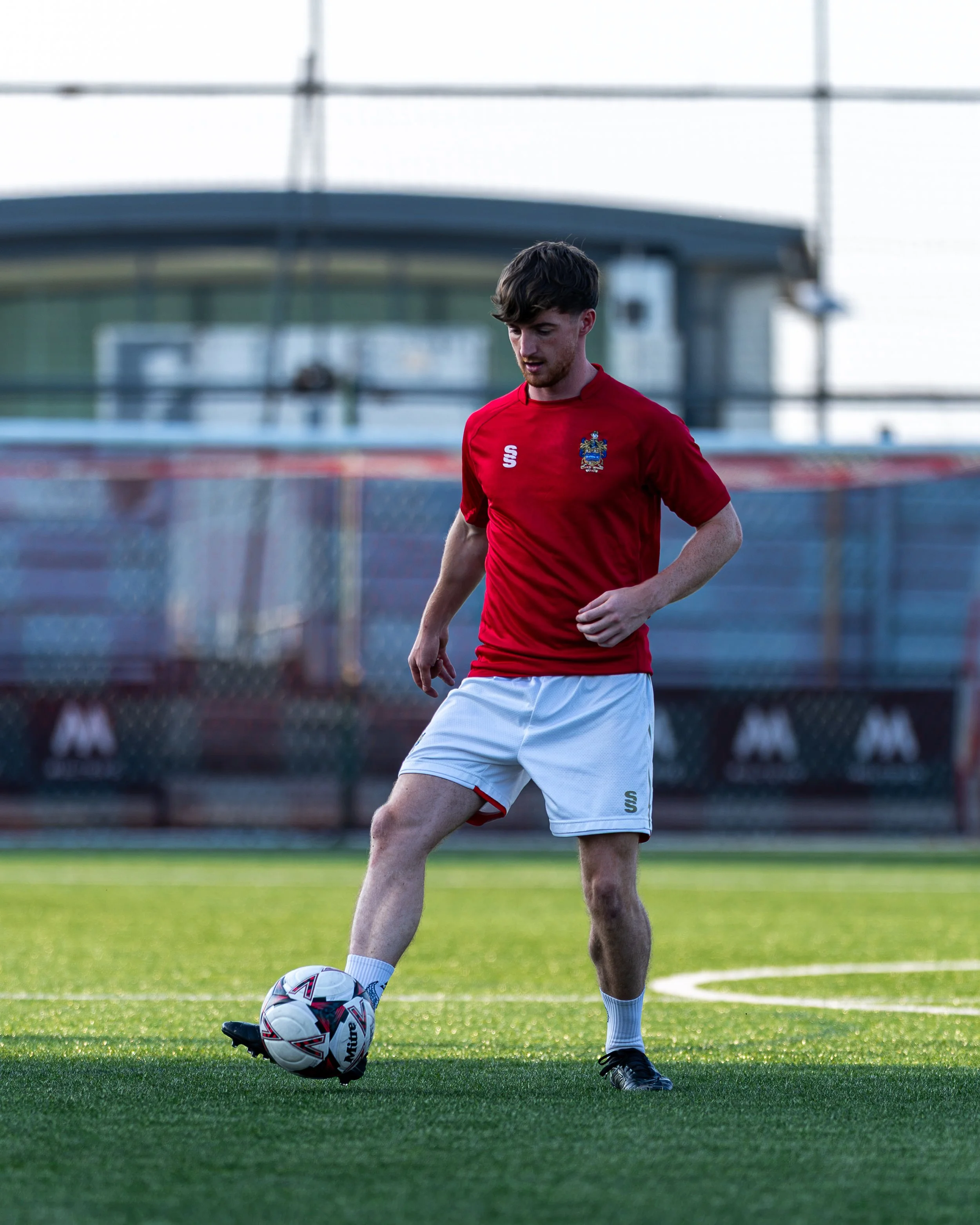 A young man in a red sports jersey and white shorts kicking a soccer ball on a grass field during daytime.