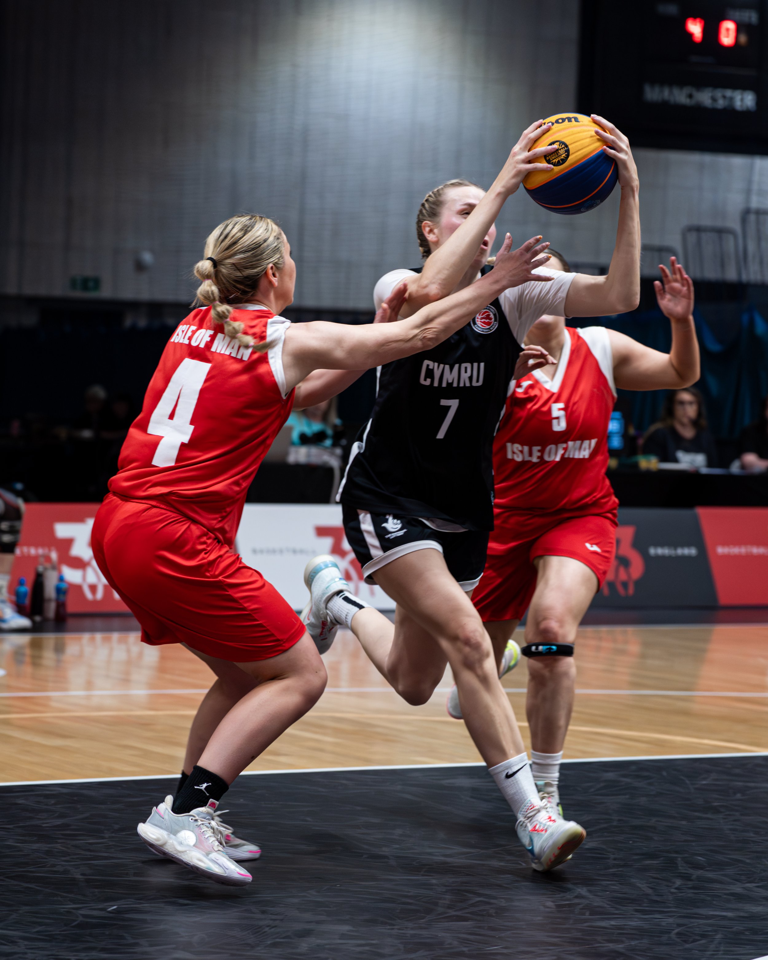Two female basketball players in action during a game, one in a red jersey and the other in a black jersey, with a gymnasium background and a scoreboard showing 40 seconds.