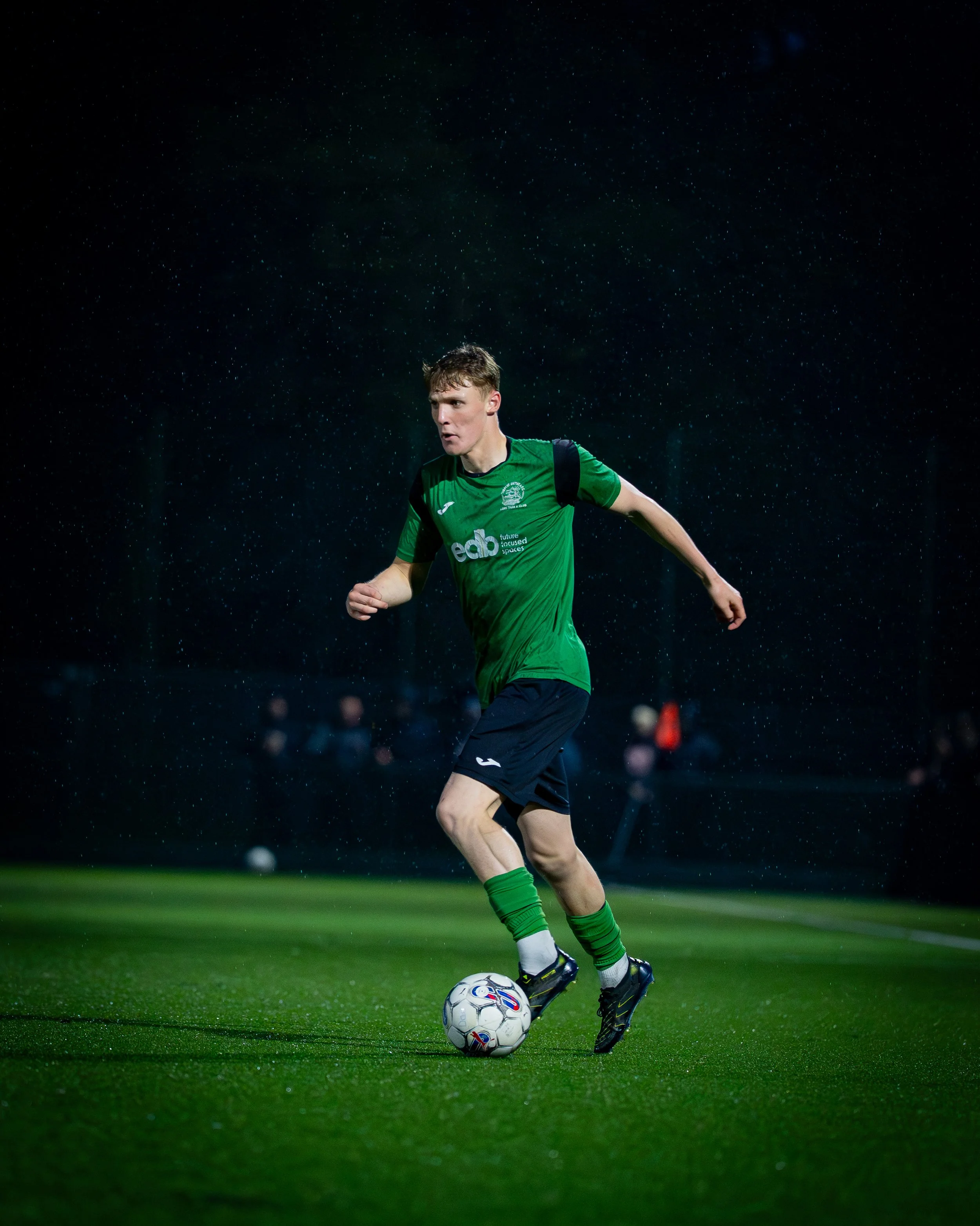 A soccer player in a green uniform kicking a soccer ball on a wet, rain-drenched field at night.
