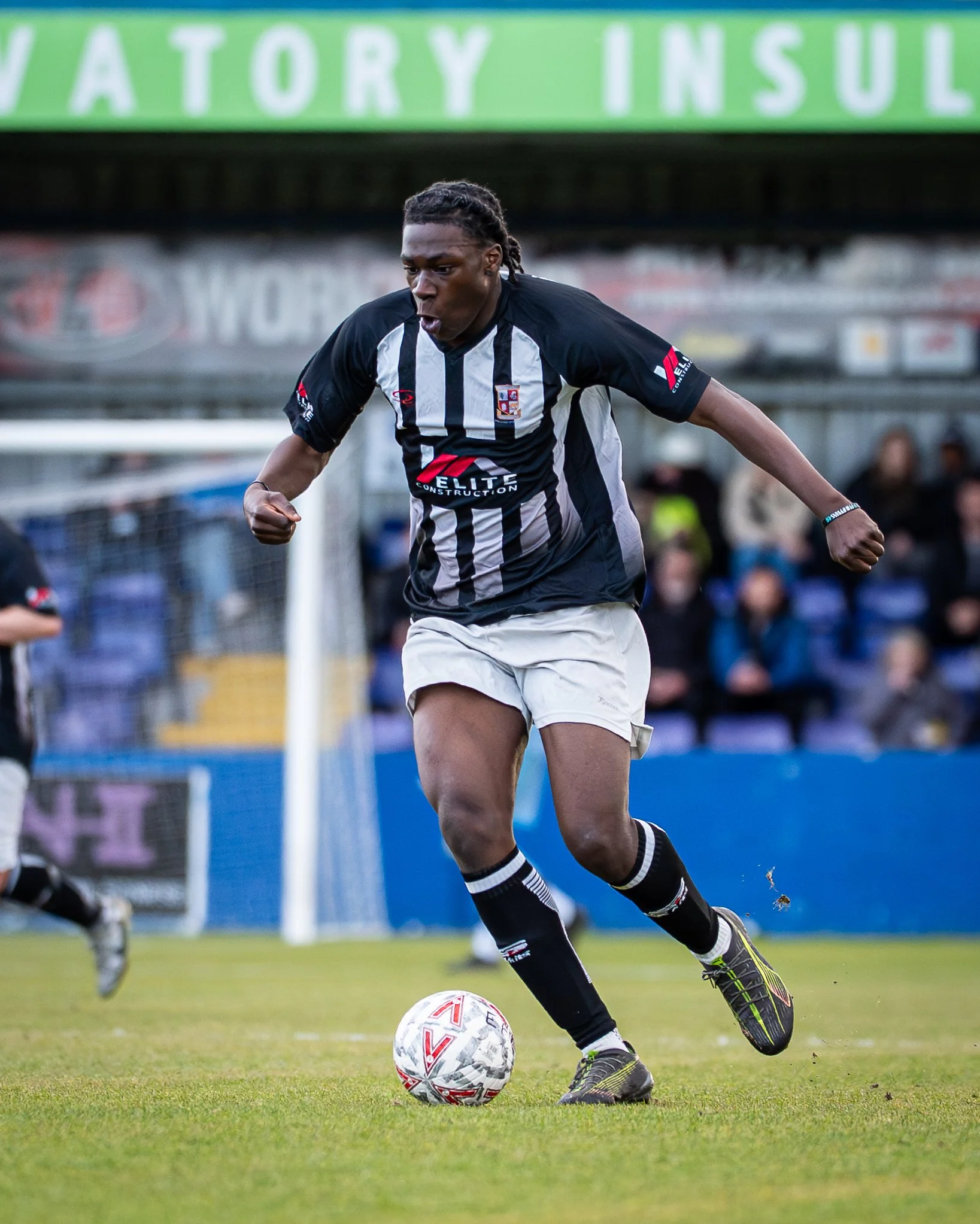 A soccer player in a black and white striped jersey, white shorts, and black socks is dribbling a soccer ball on a field. The player has braided hair, appears focused, and is wearing black athletic shoes with green accents. The background shows some 