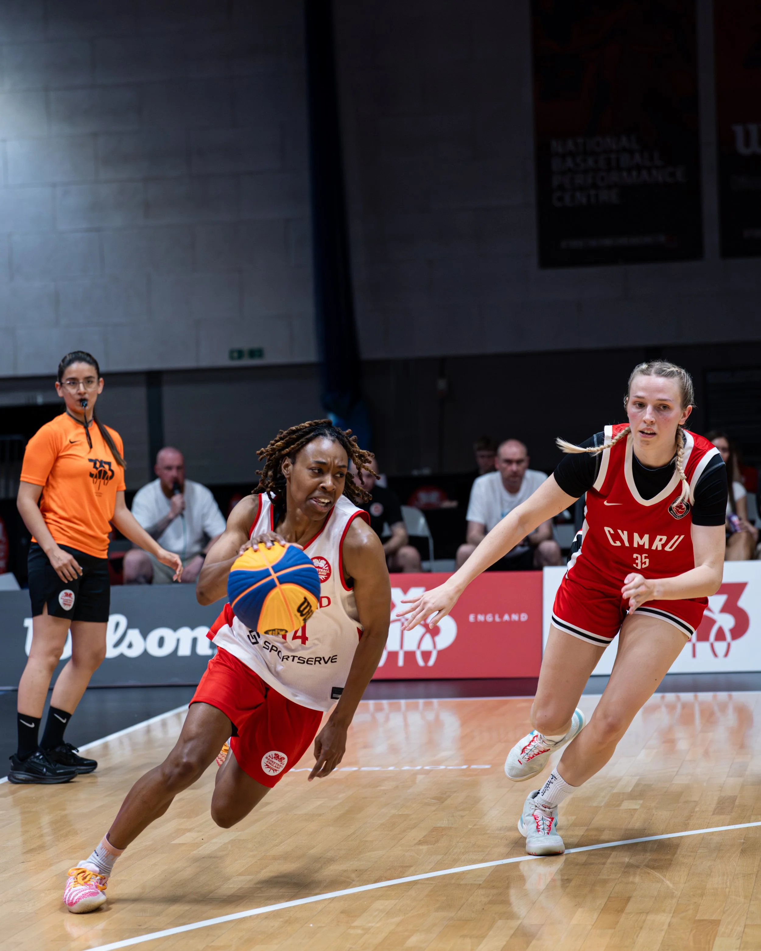 Two female basketball players competing for the ball during a game at the National Basketball Respect Performance Center. One player in a red jersey is reaching for the ball, while another player in a white jersey is defending. A referee and audience