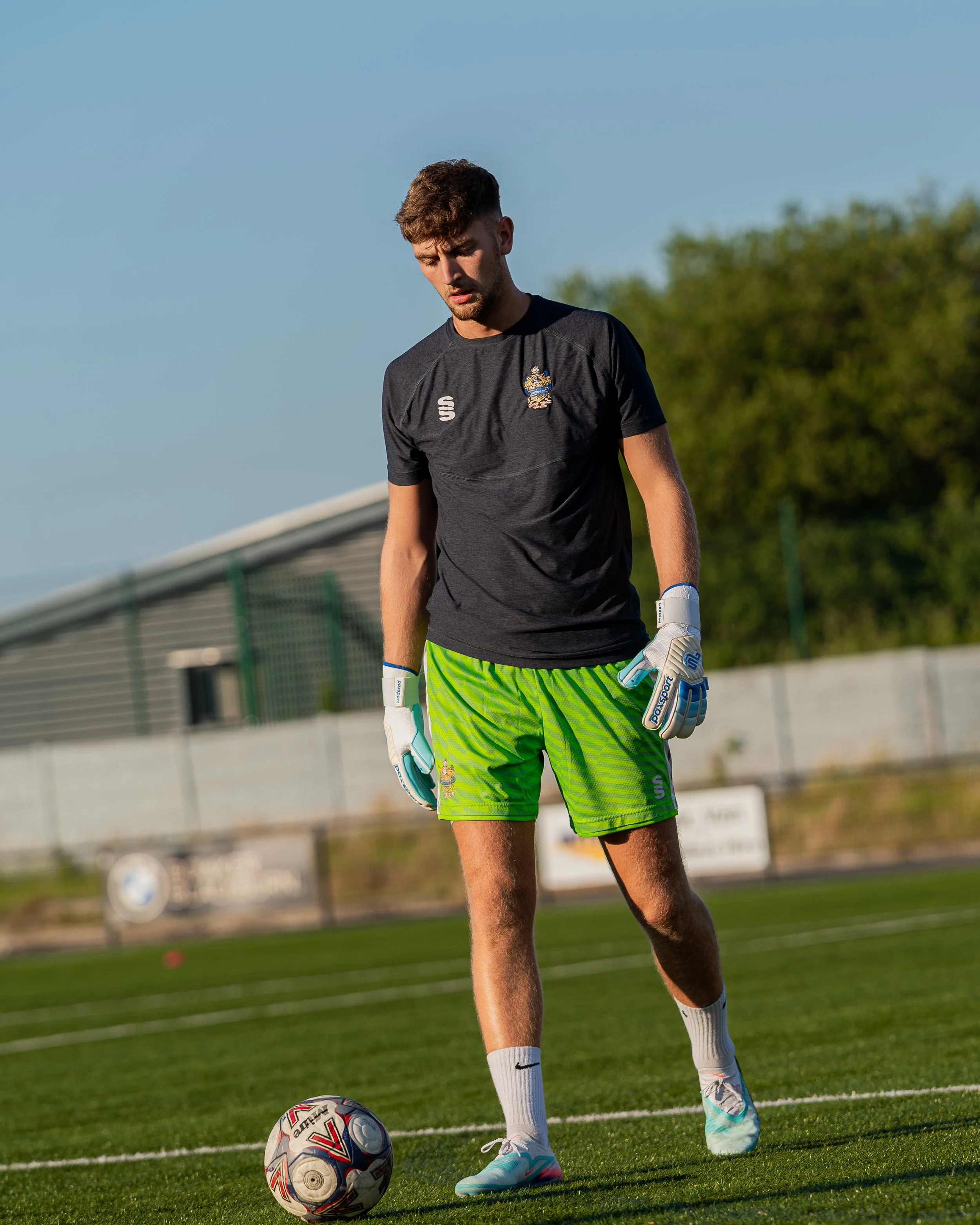 A male soccer goalkeeper wearing a black shirt, neon green shorts, white socks, and soccer cleats, standing on grass with a soccer ball at his feet during daytime.
